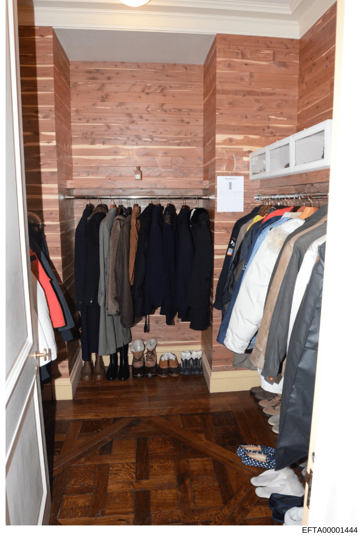 This is a photograph of a residential walk-in closet featuring wooden walls and organized clothing storage. The closet contains multiple hanging rods with jackets, coats, and outerwear in various colors (predominantly black, navy, and earth tones), a
