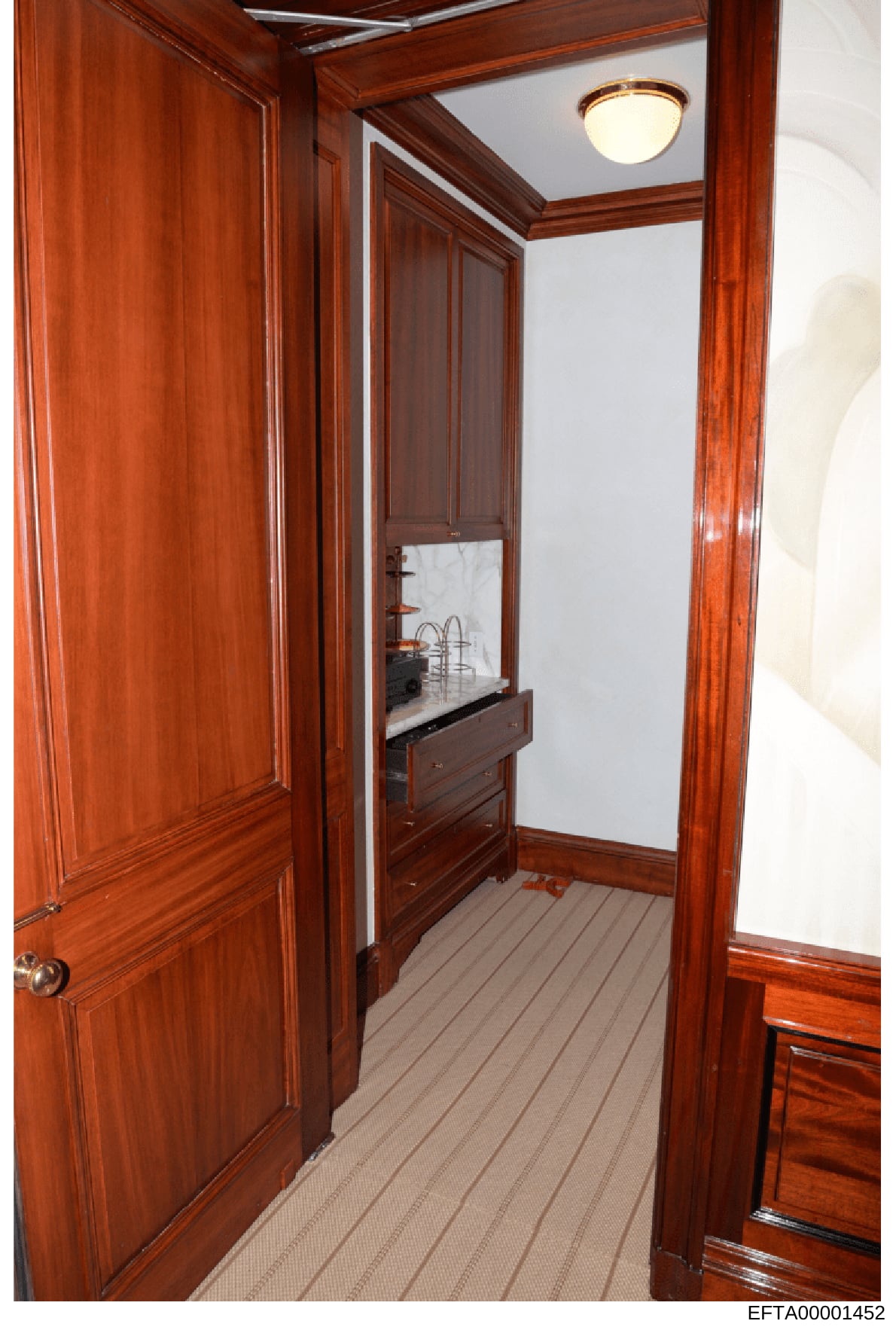 Photograph of a narrow hallway or corridor inside what appears to be a yacht or luxury vessel, featuring rich reddish-brown wood paneling throughout, a recessed sink/vanity with marble countertop, overhead lighting fixture, and carpeted flooring. The