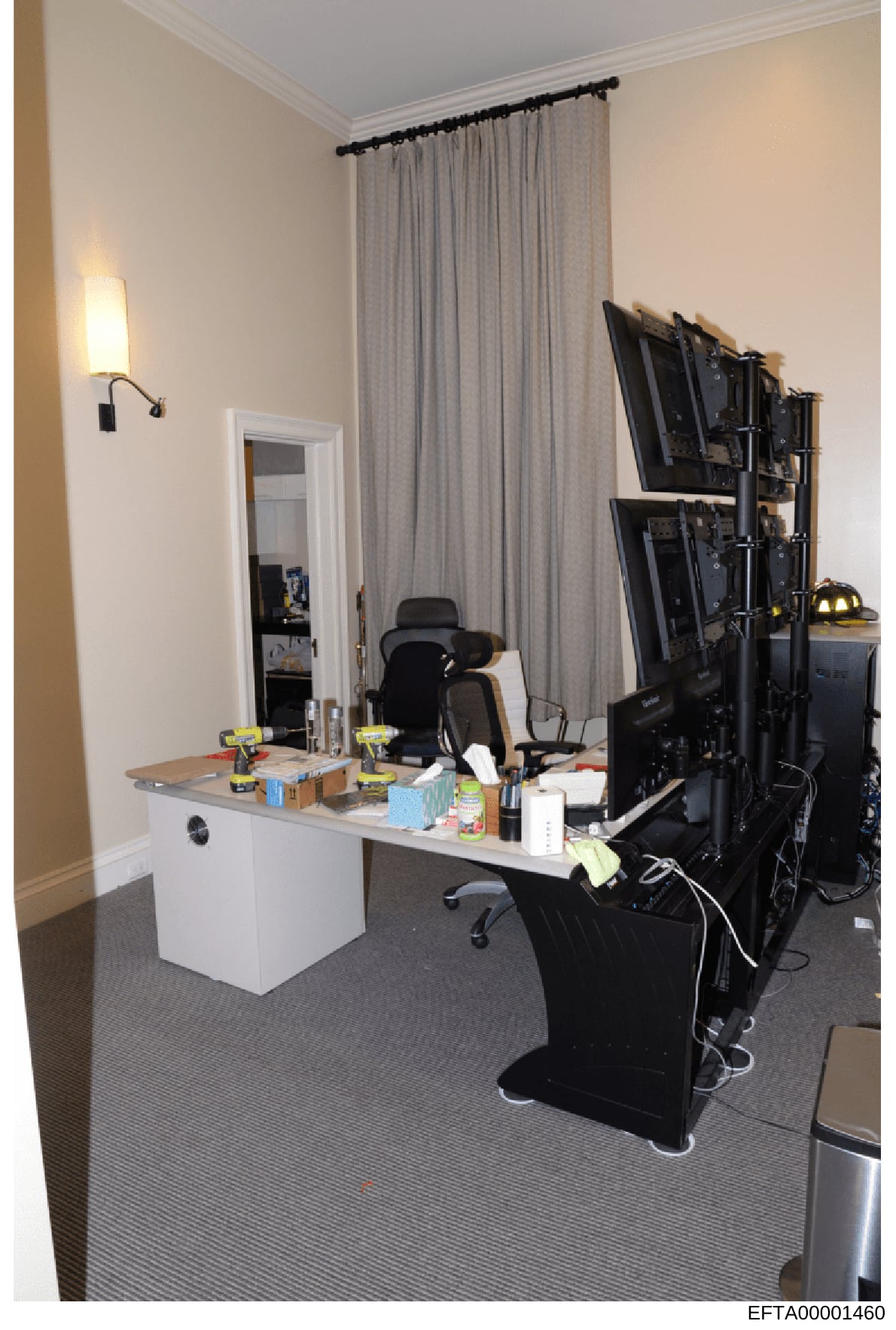 This photograph shows an interior office or workspace room containing computer equipment, desks, and furnishings. The room features a white desk with a mirror, multiple computer monitors on a stand, office chairs, and various personal items. This app