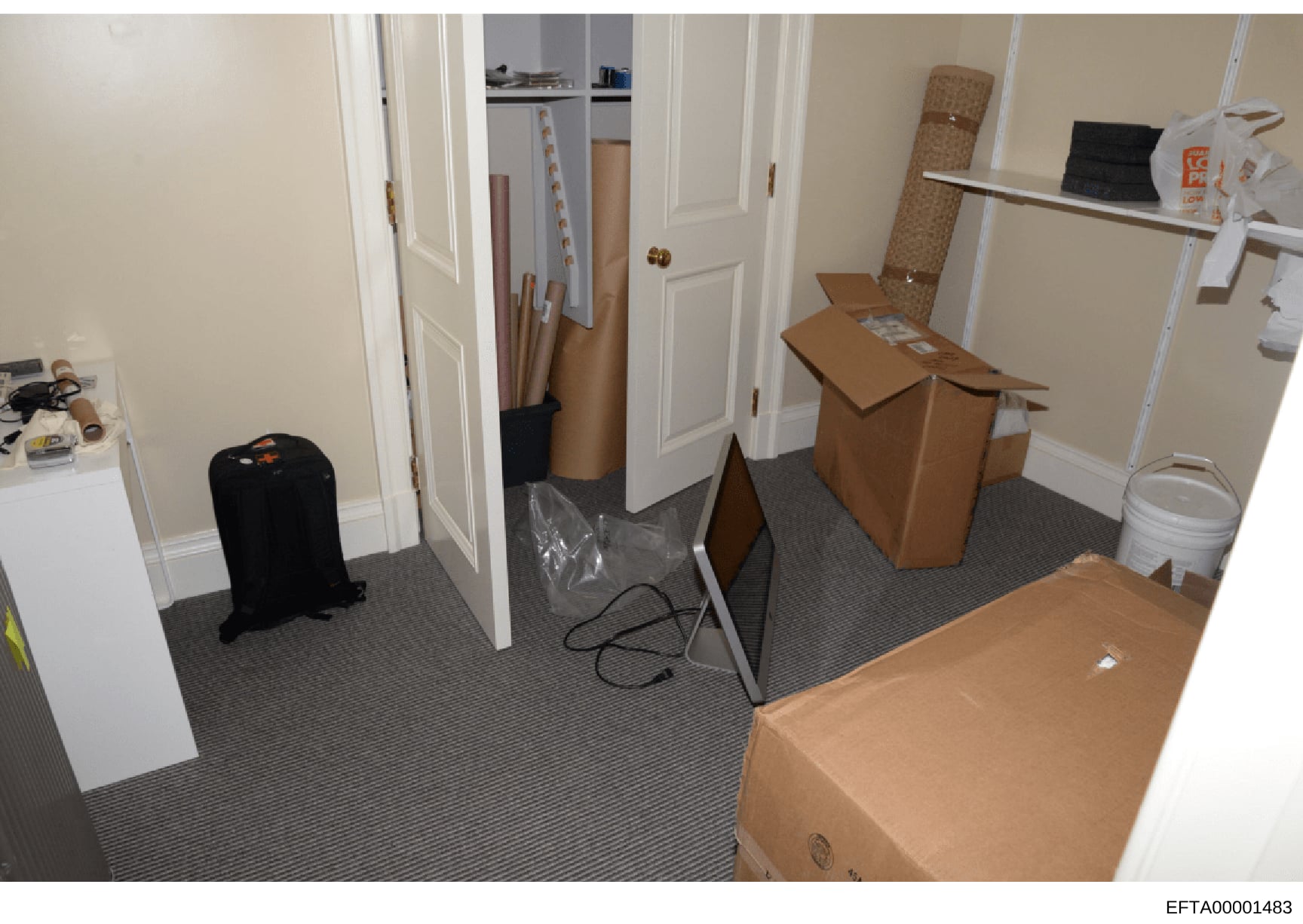 This is an evidence photograph showing an interior room containing multiple cardboard boxes, a black suitcase, rolled materials, and various storage items. The room appears to be a closet, storage area, or hallway with white doors, beige walls, and g