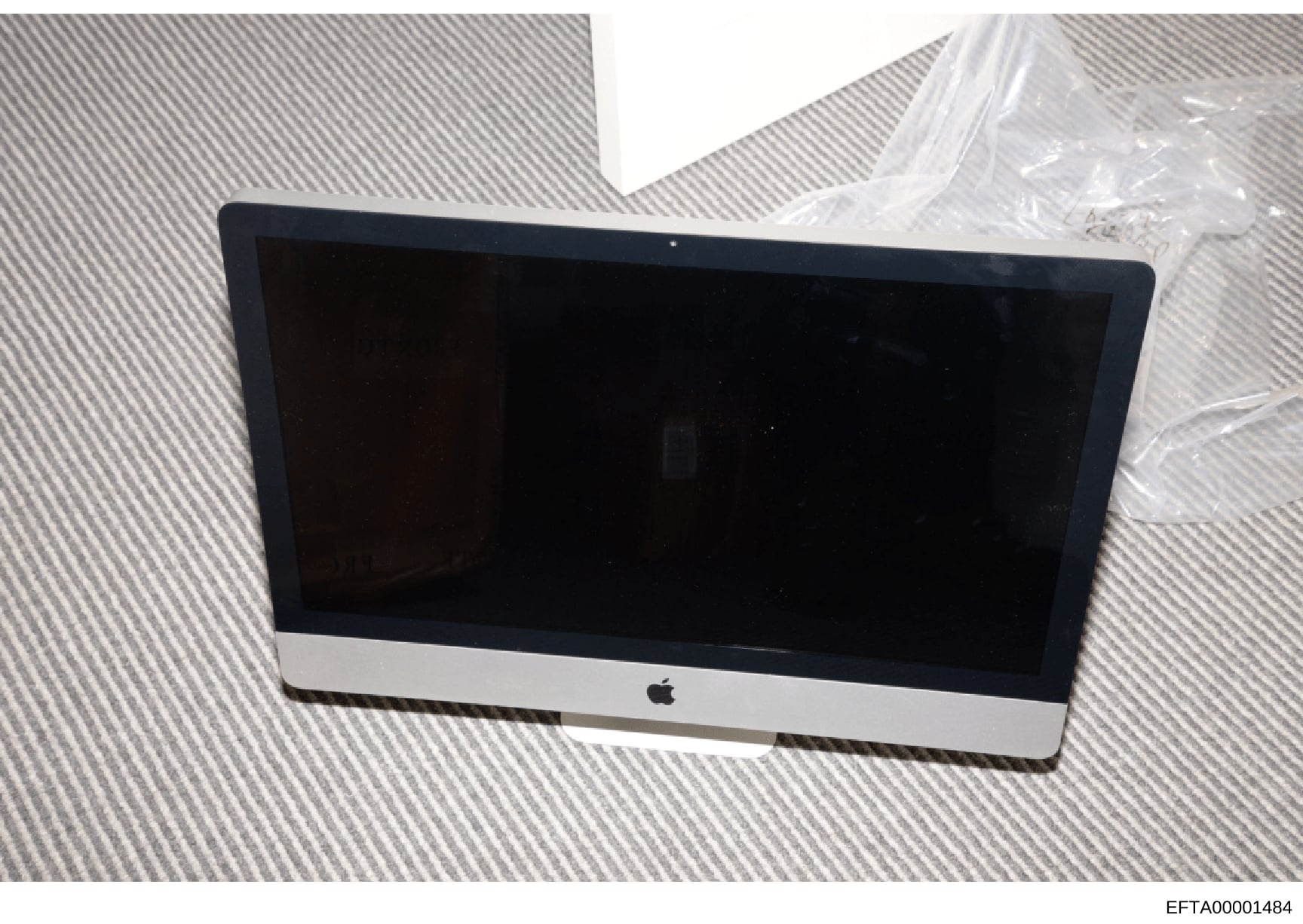 This is a photograph of a black and white Apple iMac desktop computer, likely seized as evidence during the investigation. The computer appears to be powered off or in standby mode, and is photographed on a textured surface with plastic wrapping visi