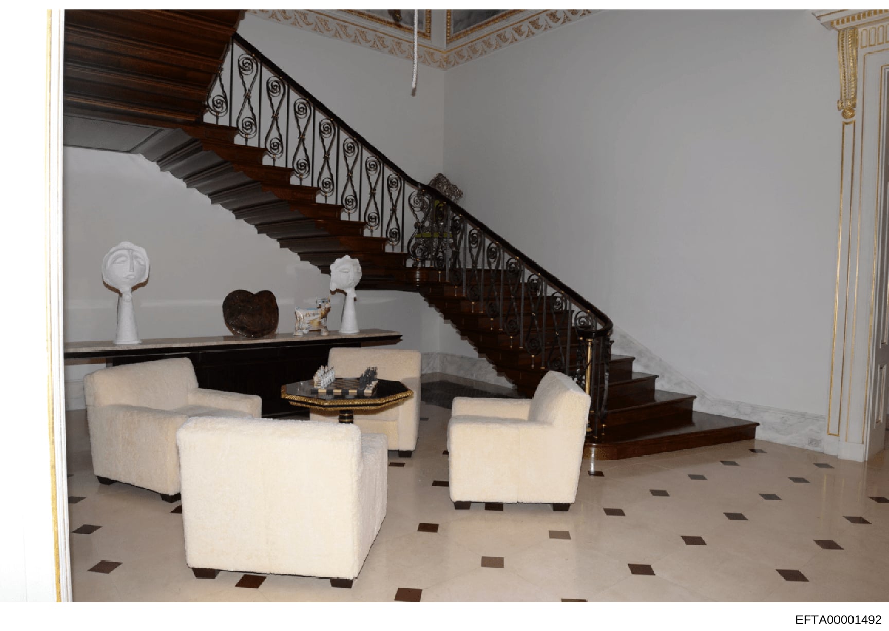 This photograph depicts the interior of a luxury residence, showing an elegant foyer with a dark wood grand staircase featuring ornate wrought iron railings, cream-colored upholstered seating furniture, decorative sculptures and art objects, and orna