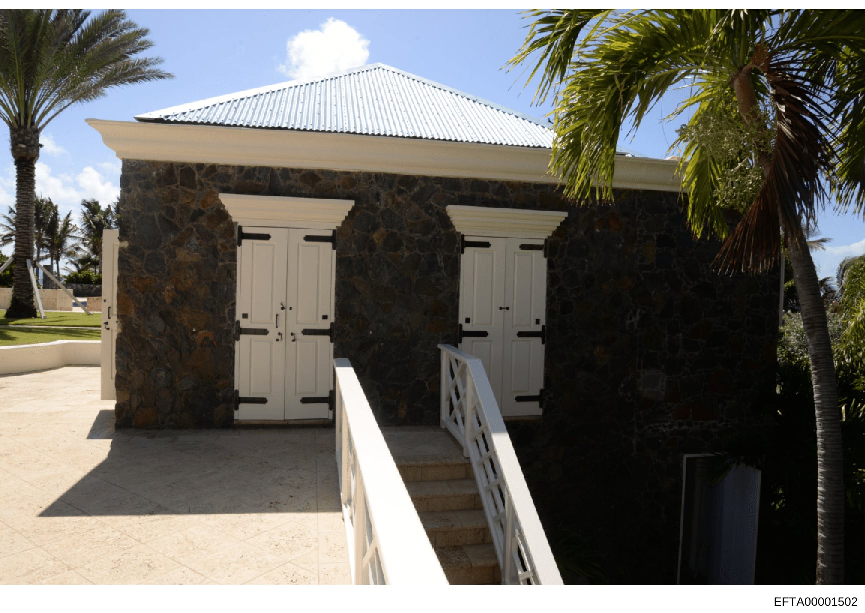 This is a color photograph of a small auxiliary building or cabana structure featuring dark stone columns, cream-colored doors, and a peaked metal roof. The building appears to be located on a tropical estate with palm trees visible in the surroundin