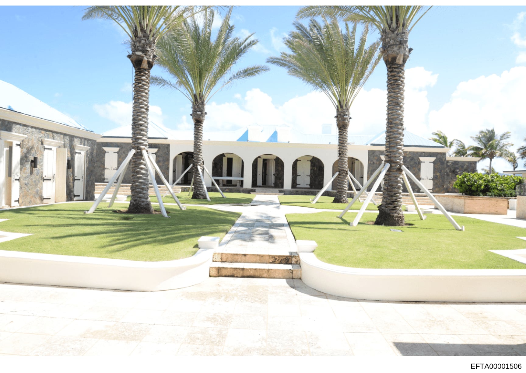 This is a photograph of the exterior courtyard and main entrance of a large residential property in a tropical location. The image shows a modern architectural design featuring multiple tall palm trees, white concrete pathways, arched architectural e