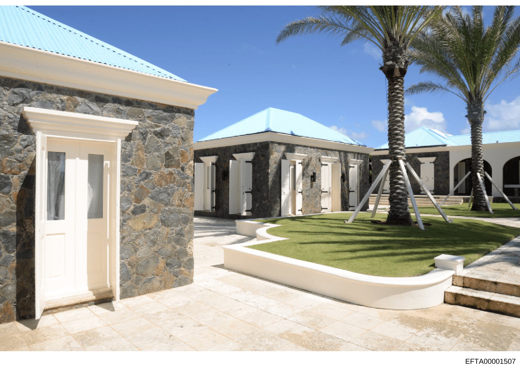 This photograph shows the exterior of a substantial residential property featuring modern stone construction with turquoise metal roofing, manicured lawns, and tropical landscaping. The image appears to be architectural documentation of an Epstein-li
