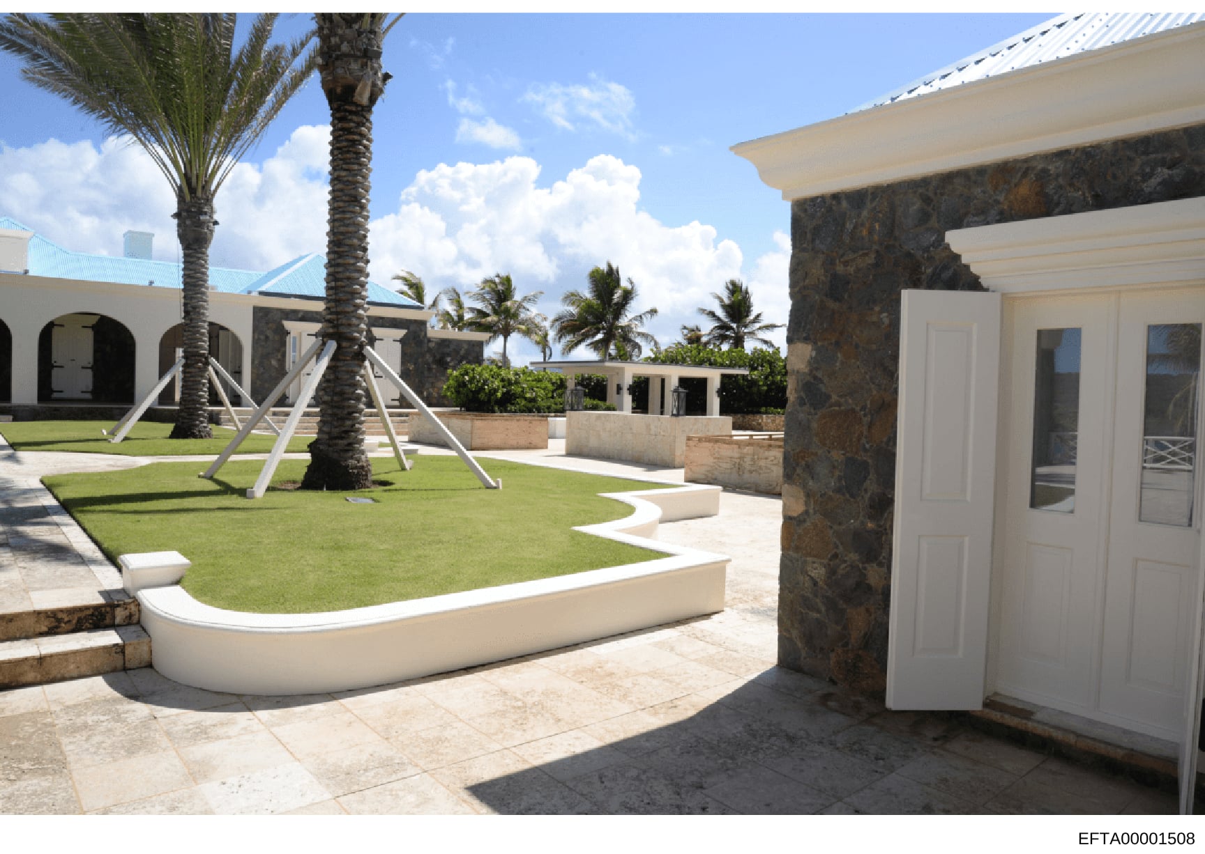 This photograph documents the exterior grounds of a high-end residential property featuring a manicured courtyard with palm trees, white architectural elements, and stone accents. The image shows the landscaping, layout, and architectural design of w
