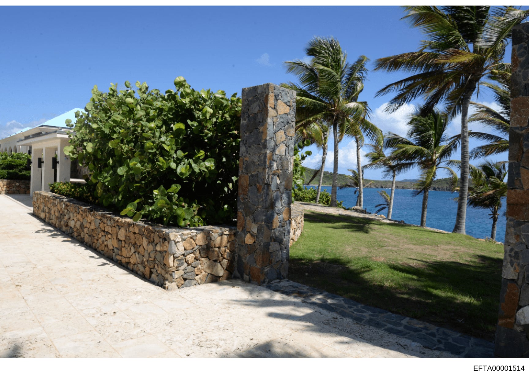 This photograph shows the exterior grounds of a tropical waterfront property, featuring a stone-constructed gateway entrance, manicured lawn, palm trees, and ocean views. The property appears to be an upscale residence with distinctive architectural 