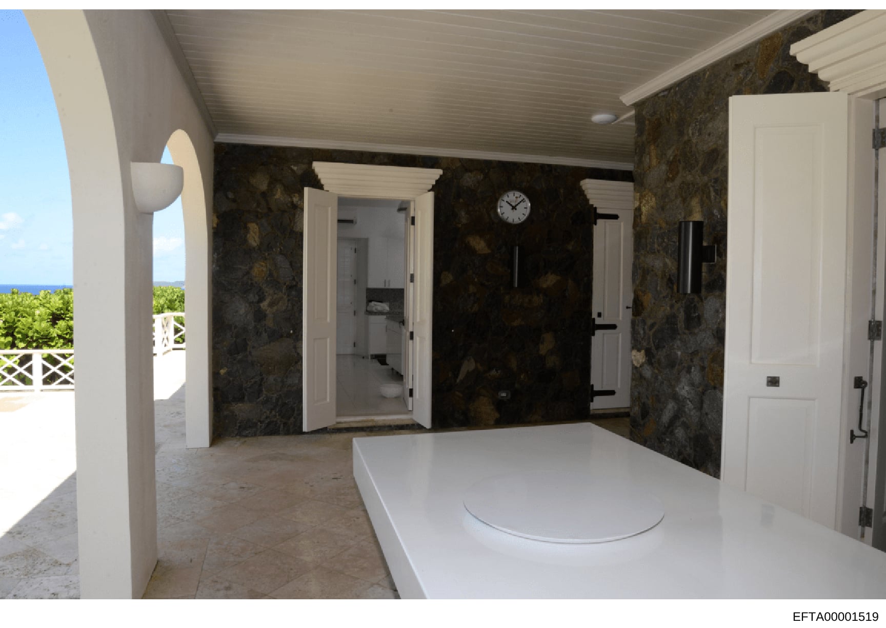 This photograph shows the interior of a luxury oceanfront property, capturing a bathroom and connecting hallway with high-end finishes including dark marble walls, white cabinetry, and ocean views visible through an open archway. The image documents 