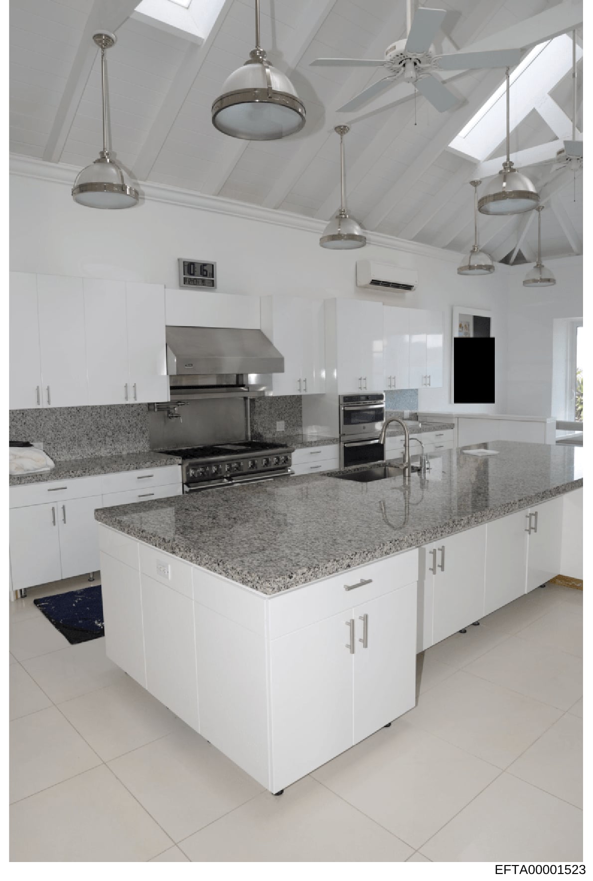 This is a photograph of a contemporary kitchen space featuring white cabinetry, granite countertops, stainless steel appliances, and professional-grade lighting. The image shows a large central island with seating area and multiple pendant lights sus