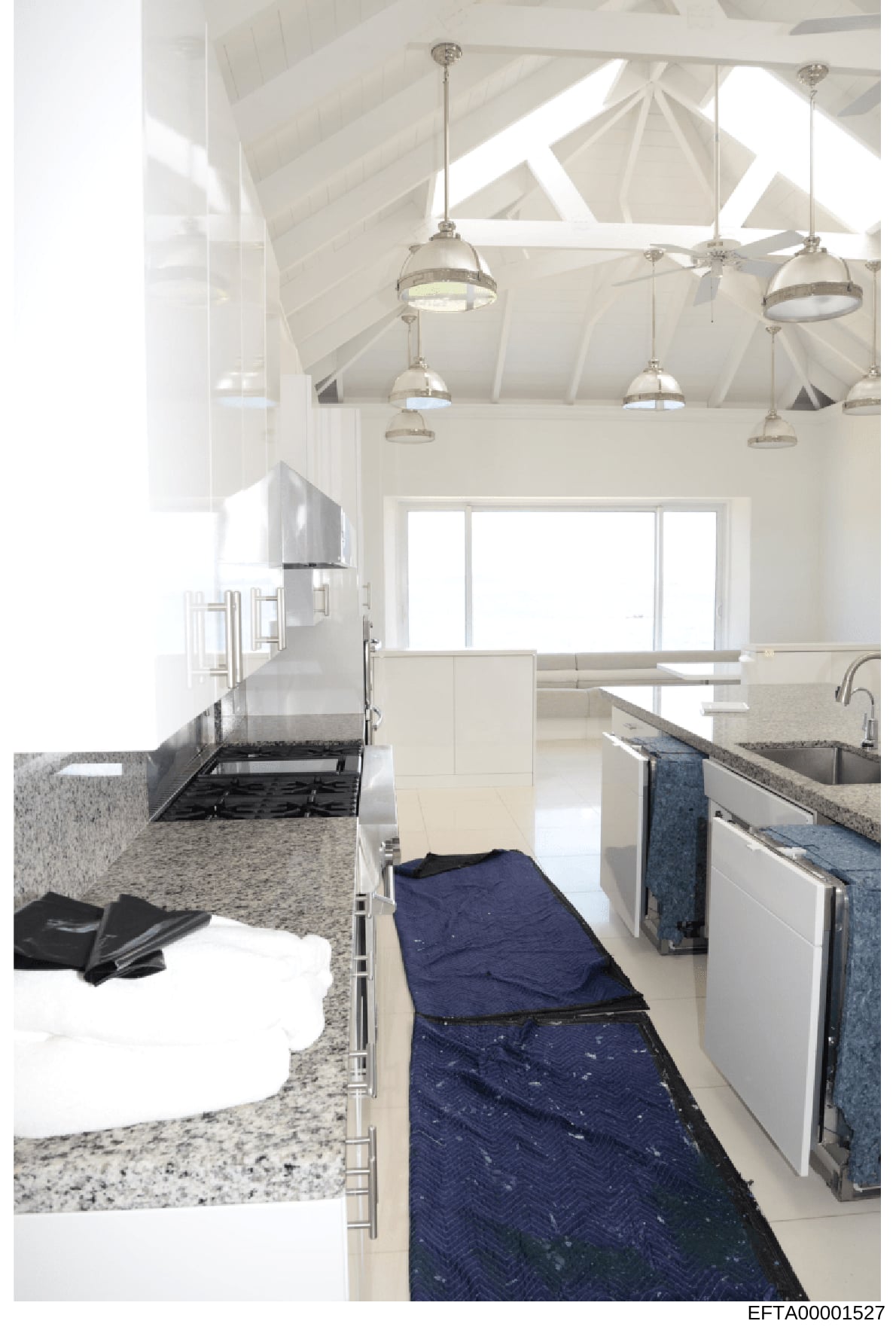This is an interior photograph of a contemporary kitchen space featuring high vaulted ceilings with exposed white beams, multiple pendant light fixtures, granite countertops, stainless steel appliances, and a blue runner rug. The image appears to be 