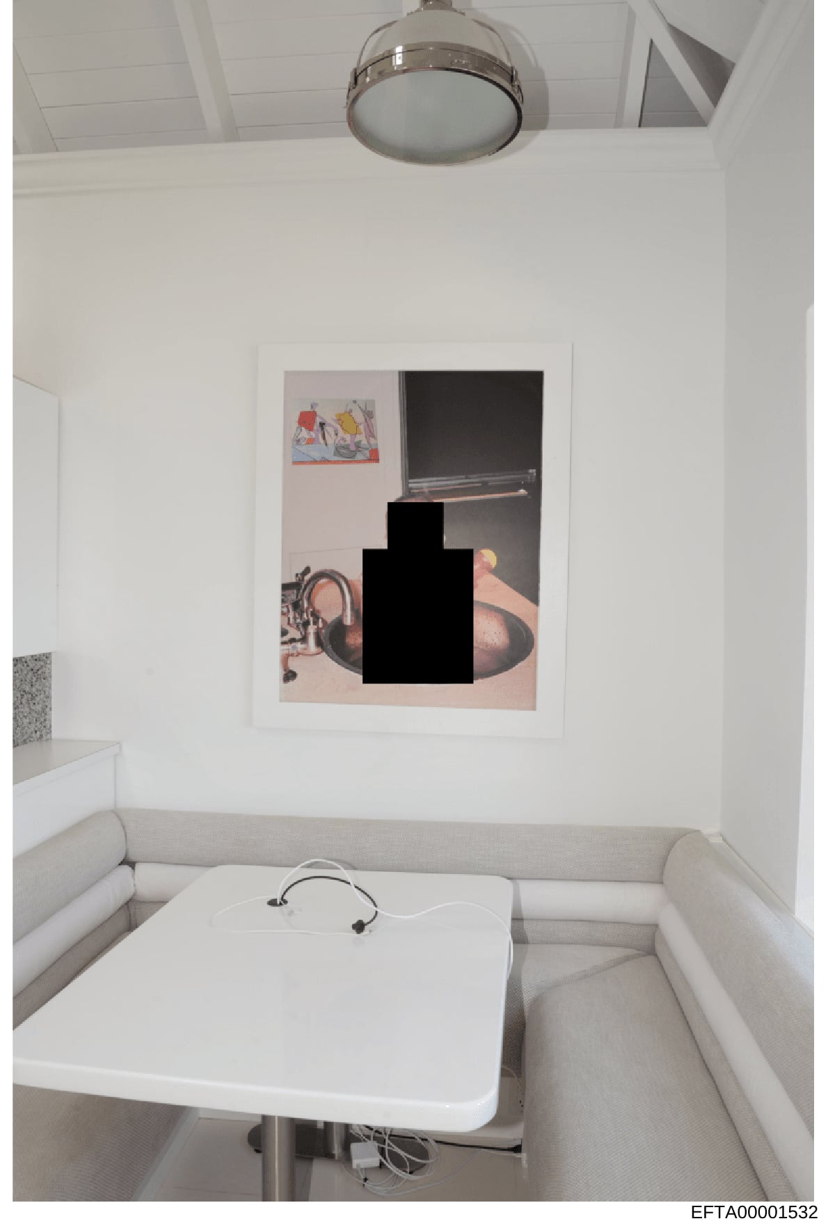 This is a photograph of a small, minimalist room with white walls and ceiling-mounted lighting. The room contains a white table or platform with headphones, beige seating along the walls, and a framed artwork on the wall with a large redacted black s