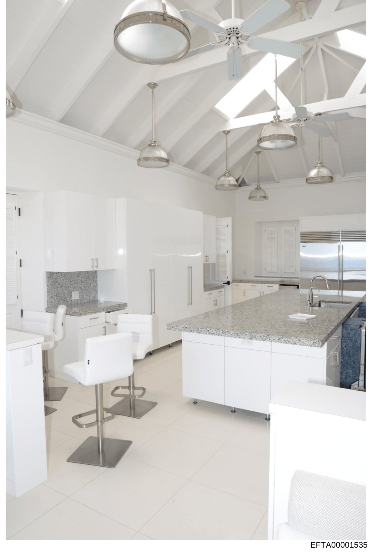 This is a photograph of a luxury kitchen interior, featuring a contemporary design with high vaulted ceilings, multiple pendant lighting fixtures, white cabinetry, granite countertops, stainless steel appliances, and an island with seating. The image