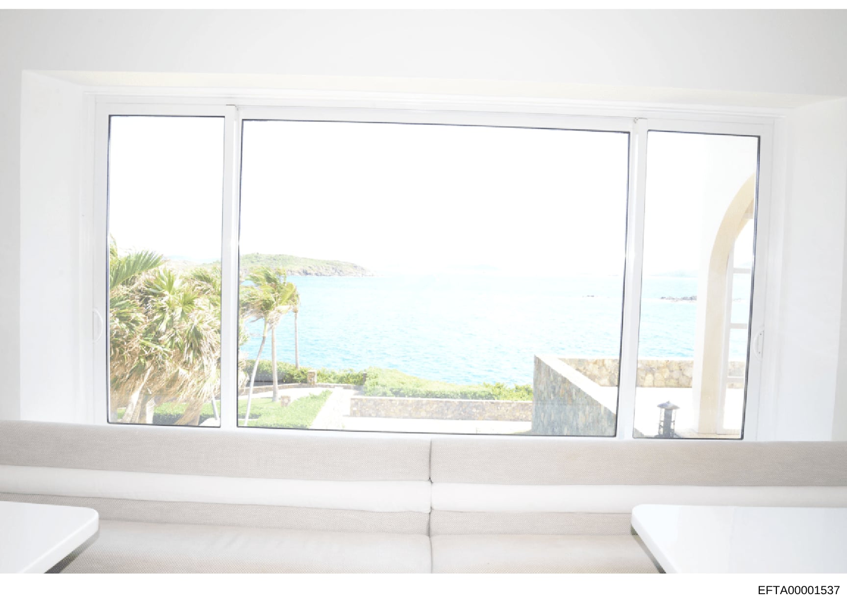 This is a photograph showing the interior of a waterfront property, taken from inside a room looking out through large floor-to-ceiling windows. The image captures a scenic coastal view with palm trees, ocean water, and distant shoreline, and appears