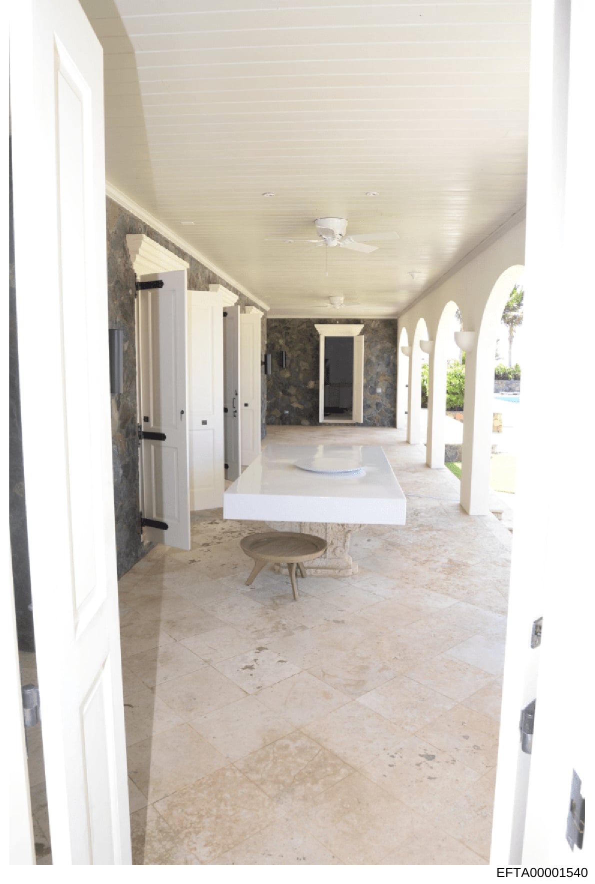 This is a photograph of an upscale residential interior space, showing a covered outdoor or semi-outdoor area with white columns, stone flooring, white cabinetry, and modern finishes. The space appears to be a loggia, pavilion, or pool house area fea