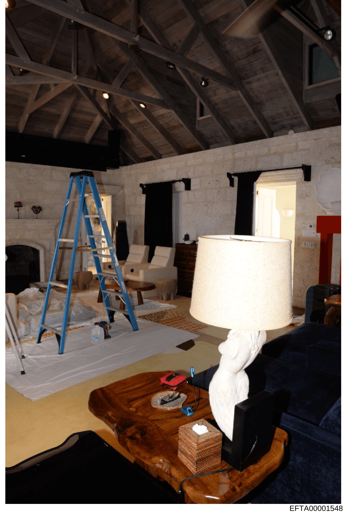 This photograph shows a spacious interior room under renovation, featuring exposed wooden beam ceiling structure, white plaster walls, and various construction materials and furniture. The image documents an active renovation project with a blue ladd