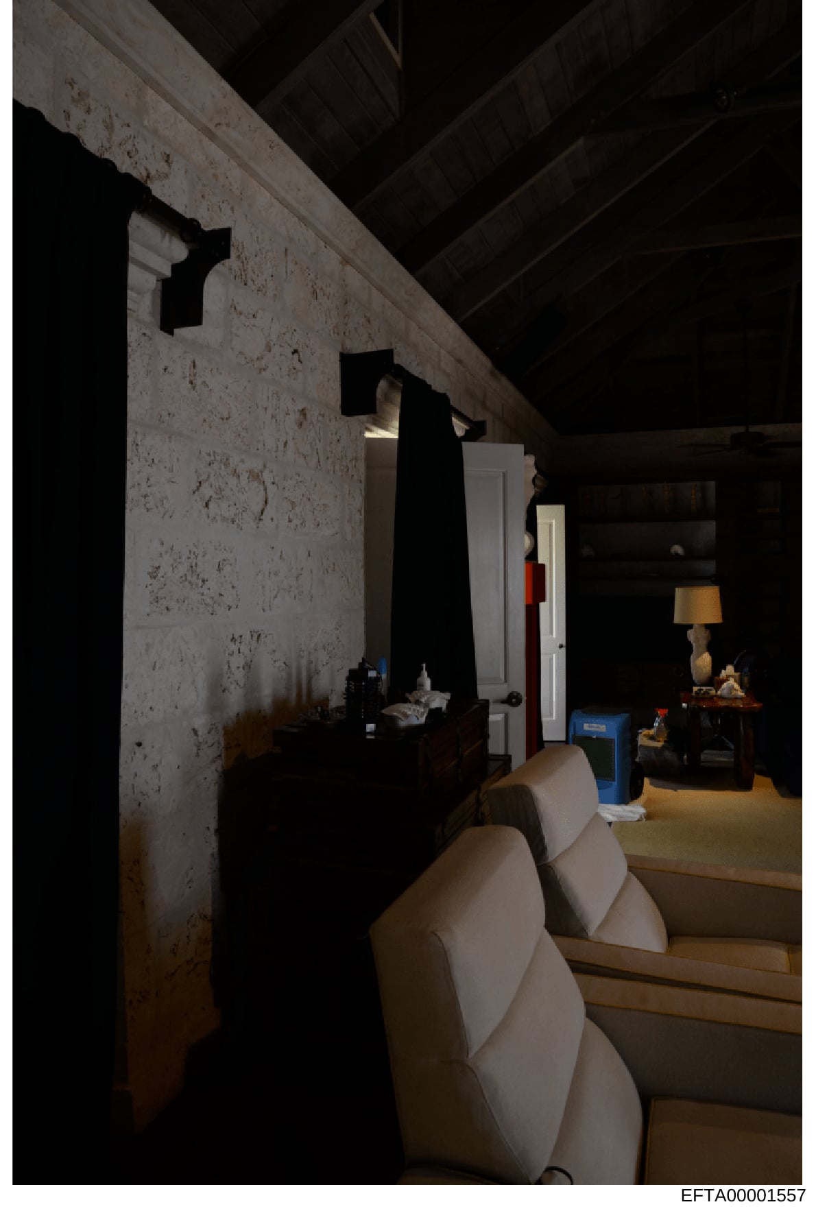This photograph documents an interior space, likely a basement or lower level room within a property under investigation. The image shows a stark, sparsely furnished room with exposed beam ceiling, concrete/plaster walls showing age and stains, white