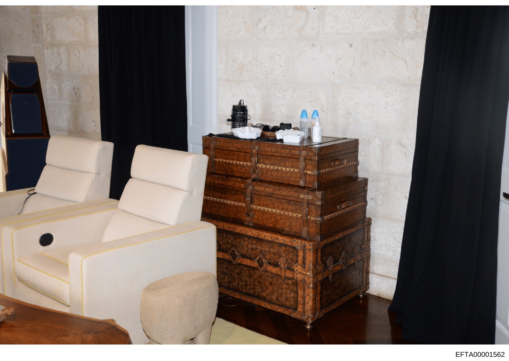 This is a photograph of an interior space showing luxury furnishings and décor, including cream-colored seating furniture with gold trim, a decorative antique wooden trunk with metal hardware, and black curtains. The room features marble or stone wal