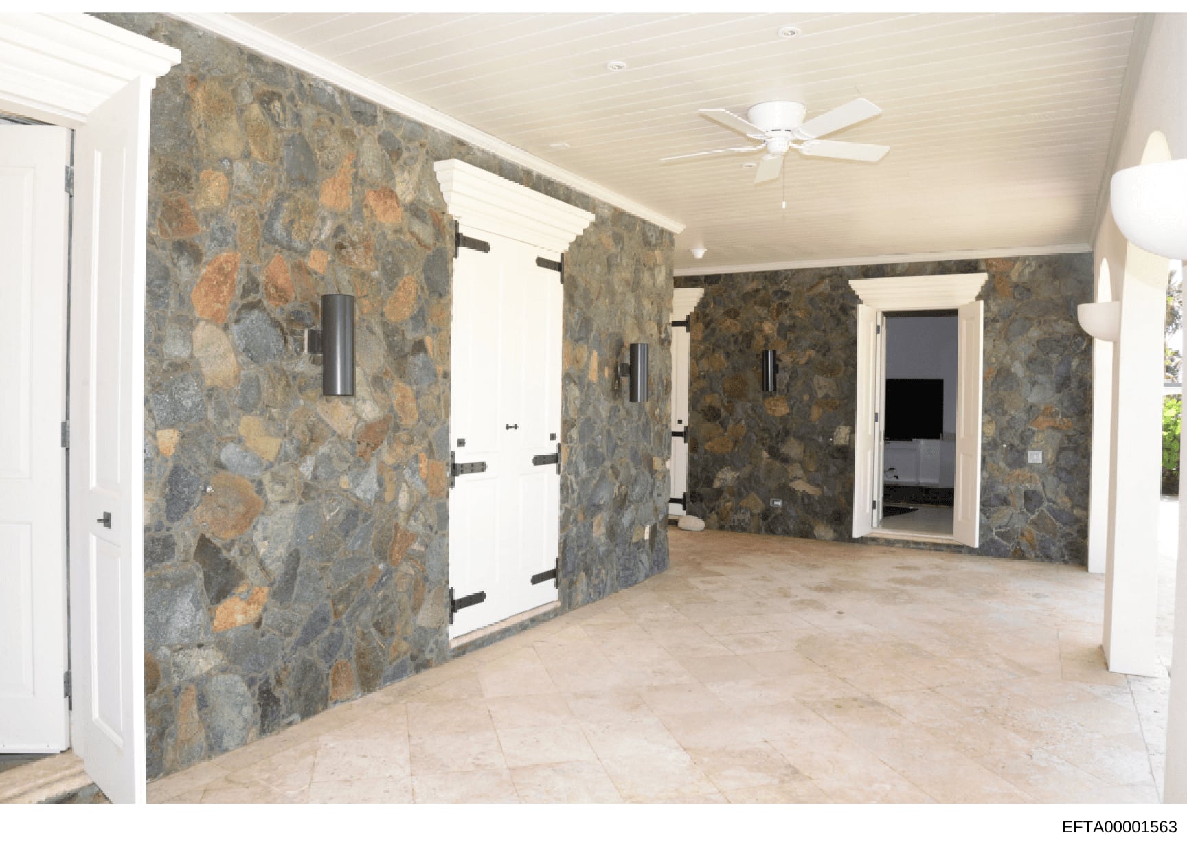 This is a photograph of a large interior room with distinctive slate or stone walls featuring earth tones and rust-colored patterns. The space shows multiple white doors with black hardware, cream-colored ceiling with ceiling fan, and light tile floo