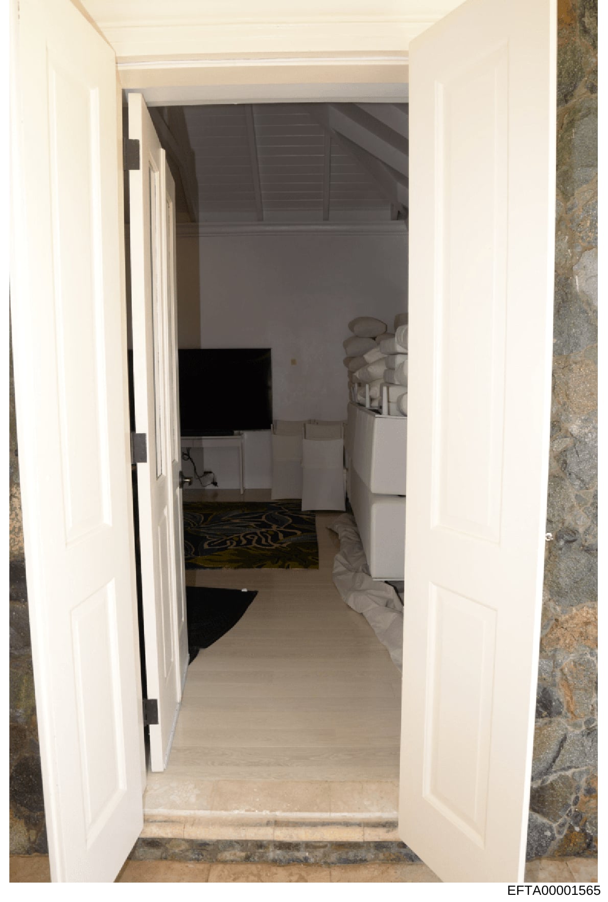 This is a photograph showing the view through an open white double door into what appears to be a bedroom or living space. The image captures the interior layout of a room with furnishings, a television, and architectural details including exposed ce