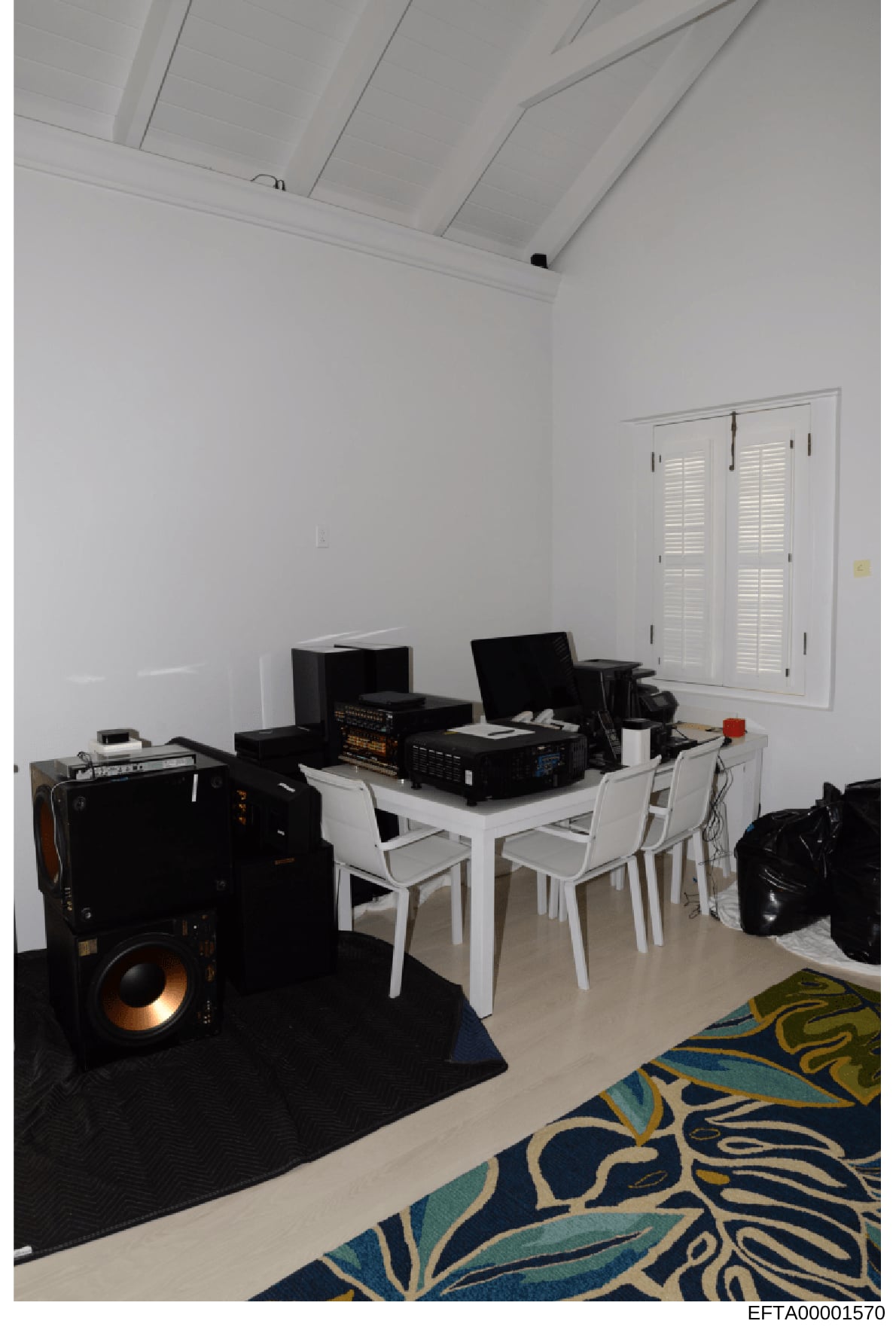 This is a photograph of an interior room containing electronic equipment, audio speakers, computer workstations, and furniture. The room features white walls, a vaulted ceiling with recessed lighting, white plantation shutters, and decorative rugs. T