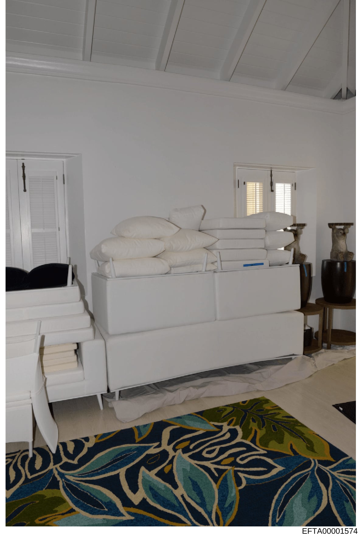 This photograph shows the interior of a bedroom featuring a white upholstered bed with pillows and folded linens, white shuttered windows, decorative wooden urns, and a patterned area rug in blue, green, and cream tones. The high white ceiling and mi