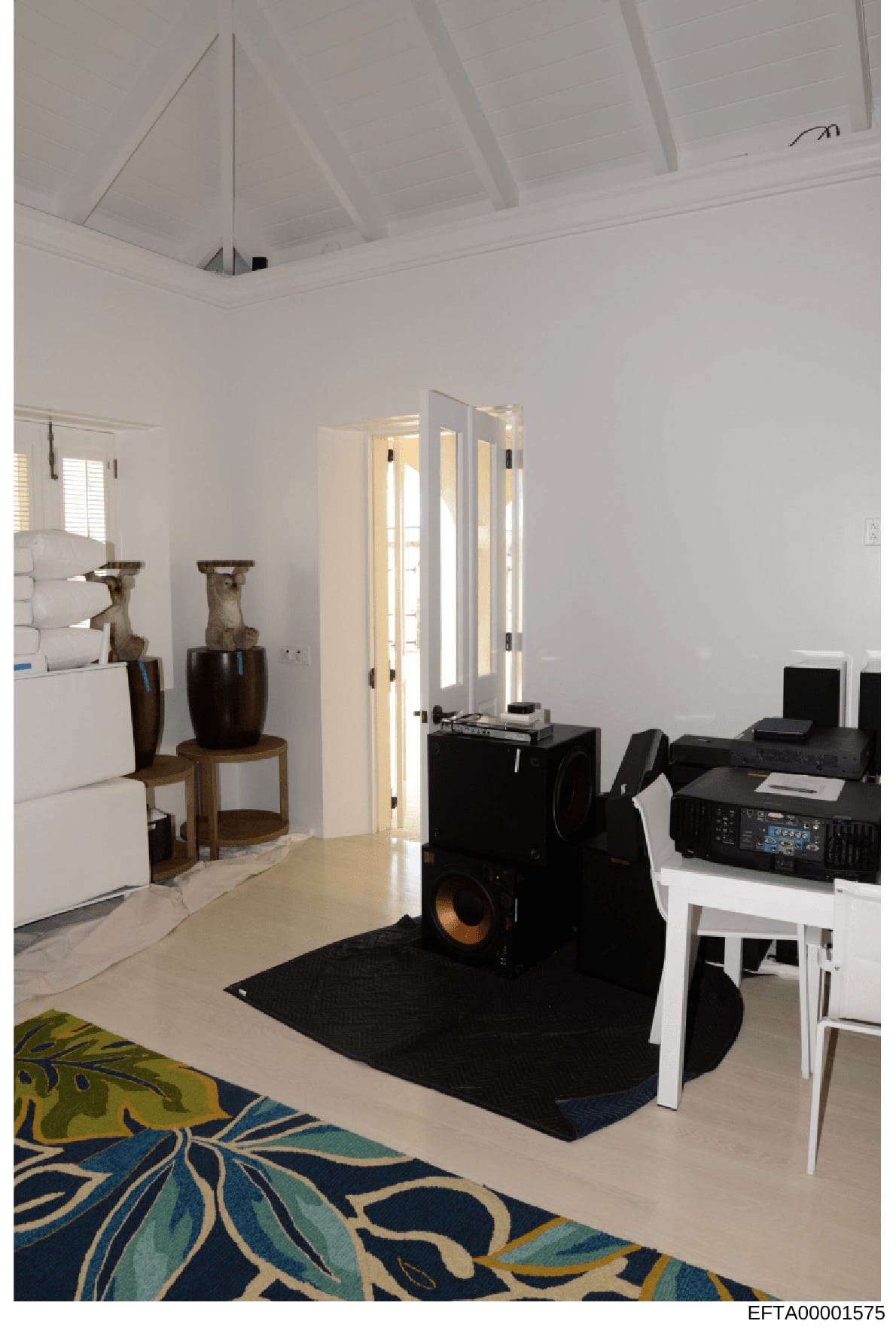 This photograph shows a contemporary interior space featuring high ceilings with exposed beams, a mix of modern and decorative furnishings including black audio/entertainment equipment, sculptural vessels, and a decorative blue and yellow area rug. T
