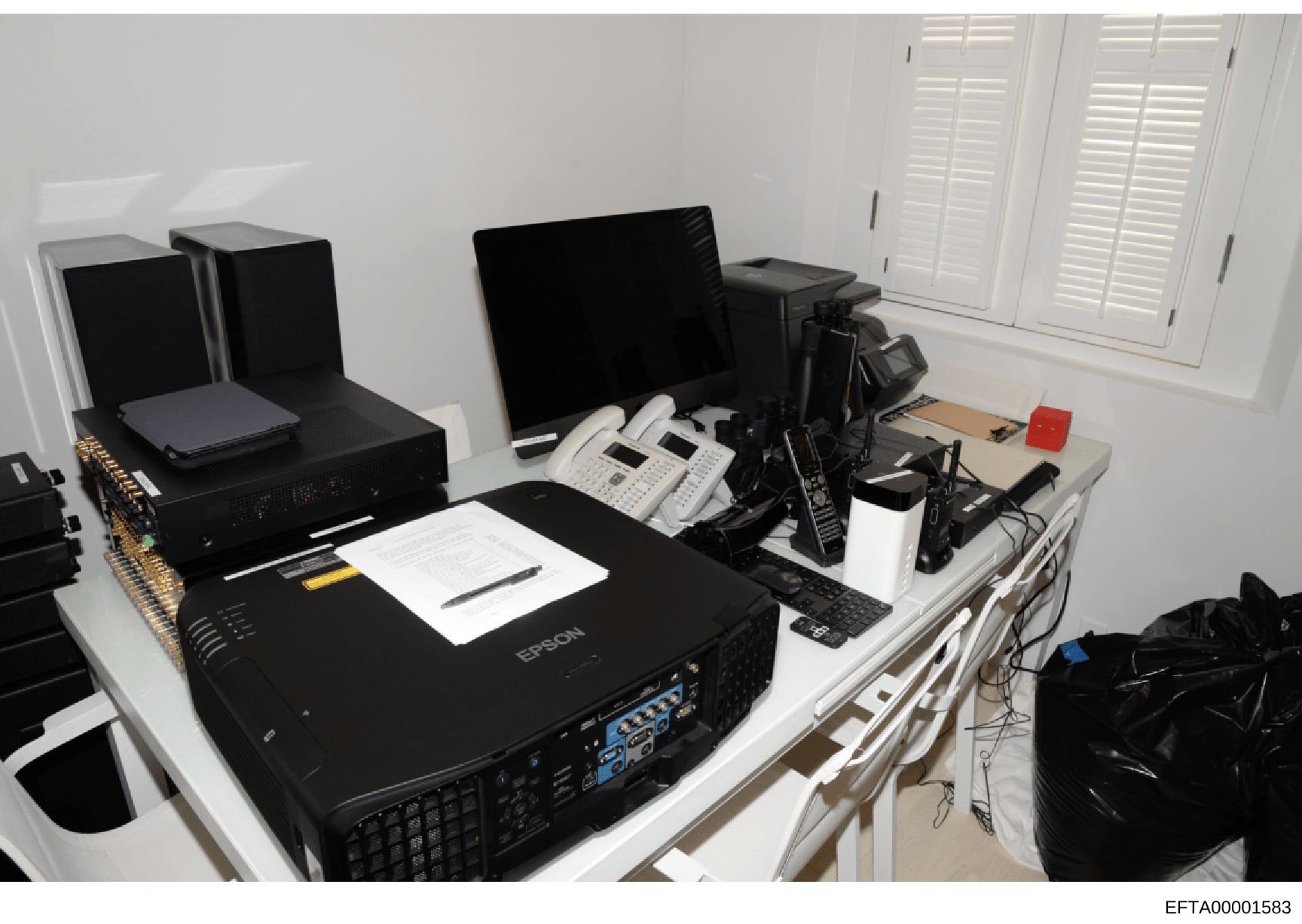 This photograph documents a computer workstation and office equipment setup, featuring multiple monitors, an Epson projector, networking equipment, white shuttered windows, and various electronic devices arranged on tables. The image appears to be ev