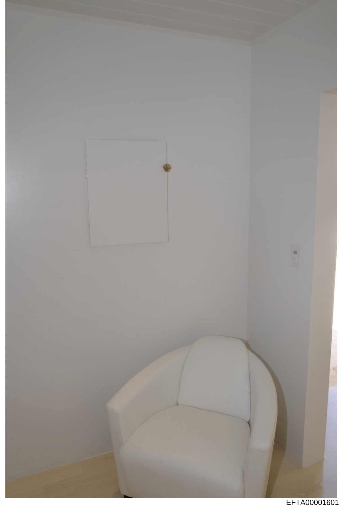 This is a photograph of an interior room space showing minimalist décor. The image depicts a white-walled room with a cream-colored upholstered armchair in the foreground and a rectangular wall-mounted panel or artwork on the wall. The photo appears 