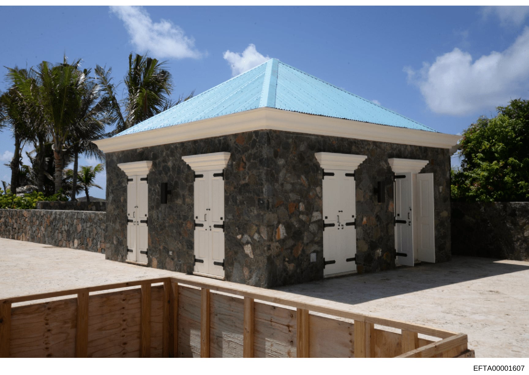 This is a photograph of a modern outdoor pavilion or shelter structure featuring a distinctive turquoise/light blue metal roof, dark stone pillars, white doors, and located in what appears to be a tropical setting with palm trees. The image shows the