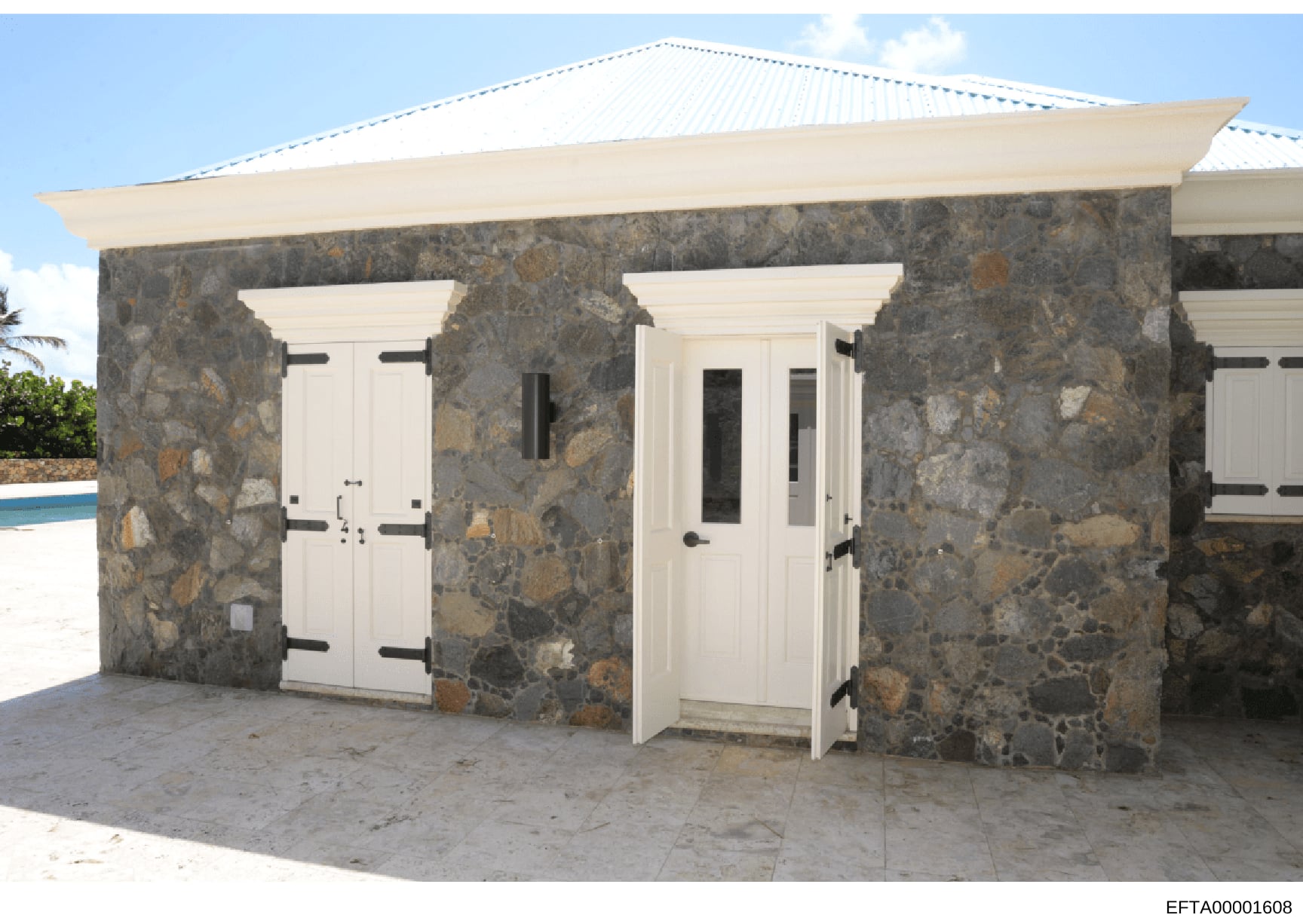 This is a photograph of a modern stone outbuilding or cottage featuring gray stone walls with white trim, white wooden doors with black hardware, and a white metal roof. The structure appears to be located on a tropical property near a swimming pool,