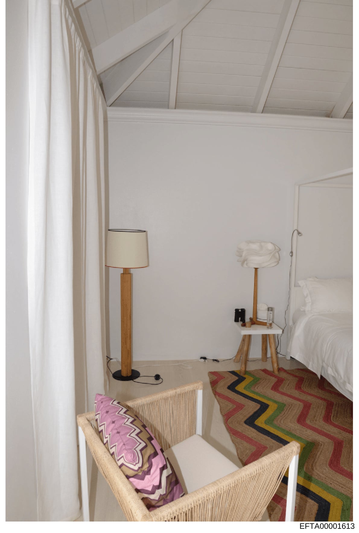 This is an evidence photograph showing a bright, minimally furnished bedroom with white walls, vaulted ceiling, and a bed with white linens. The room contains a wooden floor lamp, a side table with white pitcher, a wicker chair with a pink patterned 