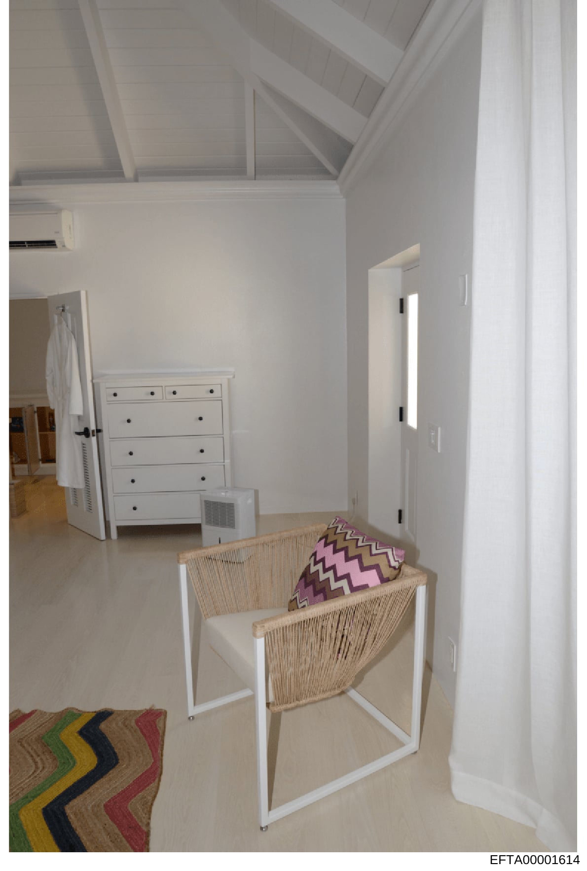 This is a photograph of a bright, minimalist bedroom interior featuring white walls, vaulted ceilings, and contemporary furnishings. The room contains a woven accent chair with decorative pillows, a white dresser, and geometric patterned area rug. Th
