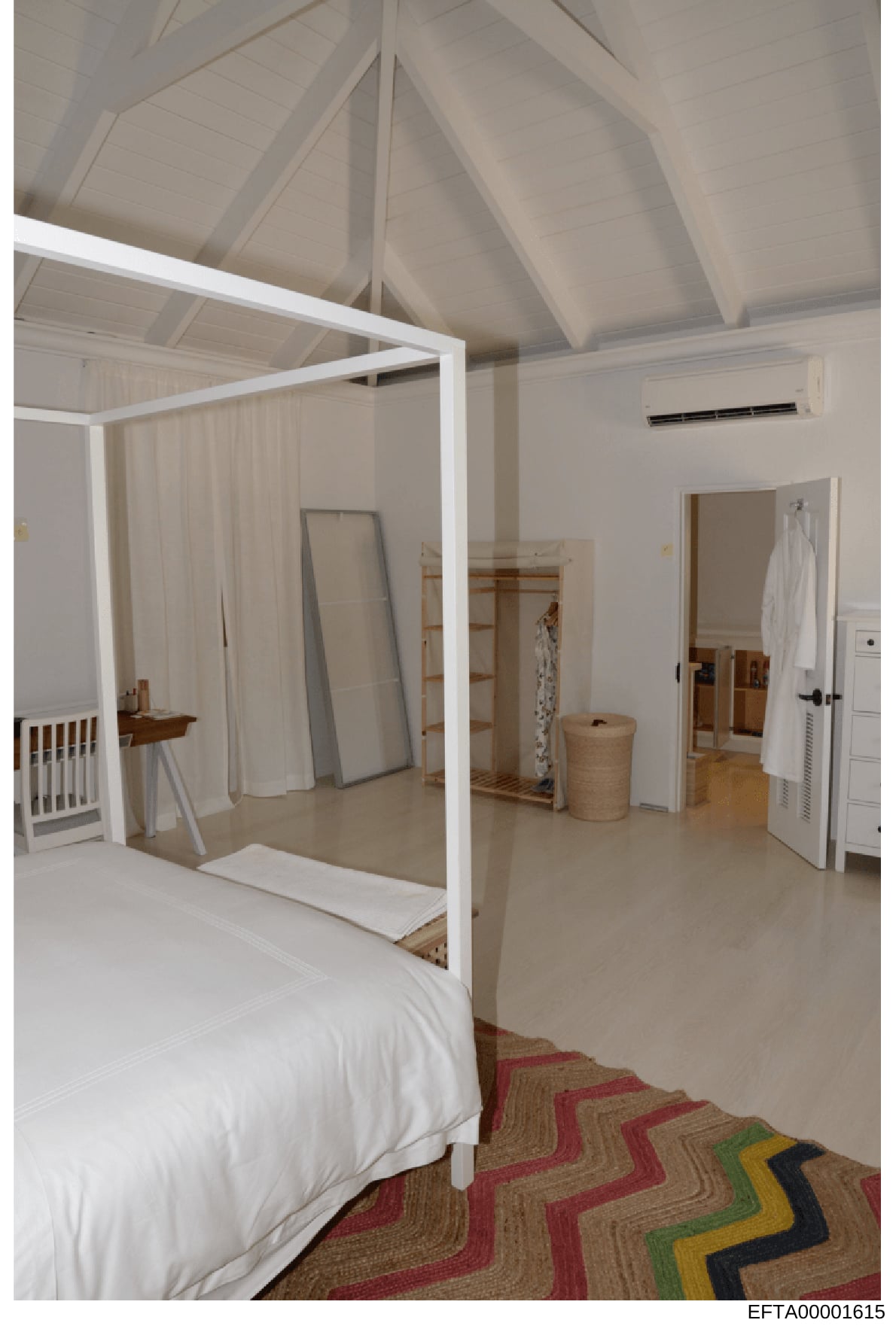 This is a photograph of a bedroom interior featuring a white four-poster bed, vaulted white ceiling with exposed beams, hardwood flooring, and a colorful geometric striped area rug in red, tan, green, and blue tones. The room appears to be part of a 