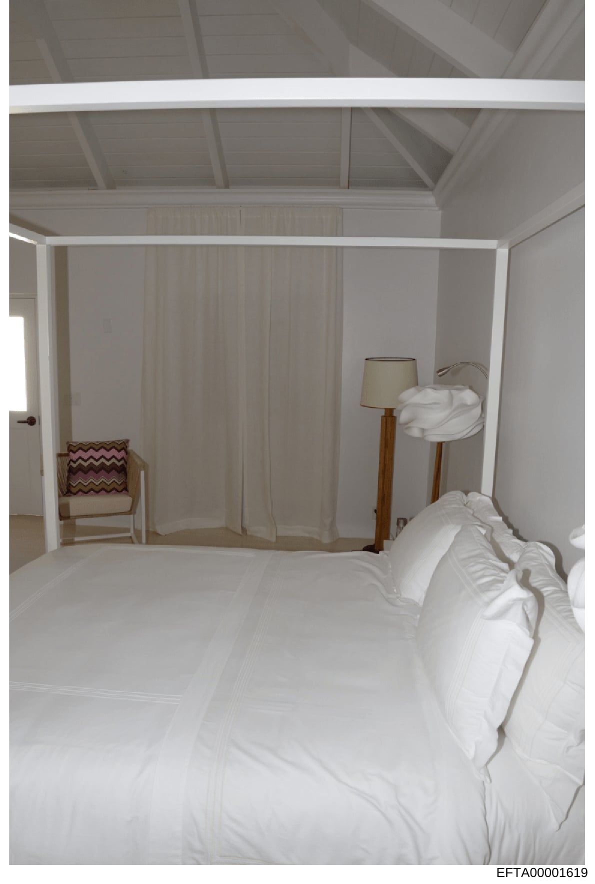 This is a photograph of an interior bedroom space, showing a four-poster bed with white linens, sheer white curtains, wooden furniture including a chair with burgundy patterned cushion, a floor lamp with beige shade, and exposed beam ceiling details.