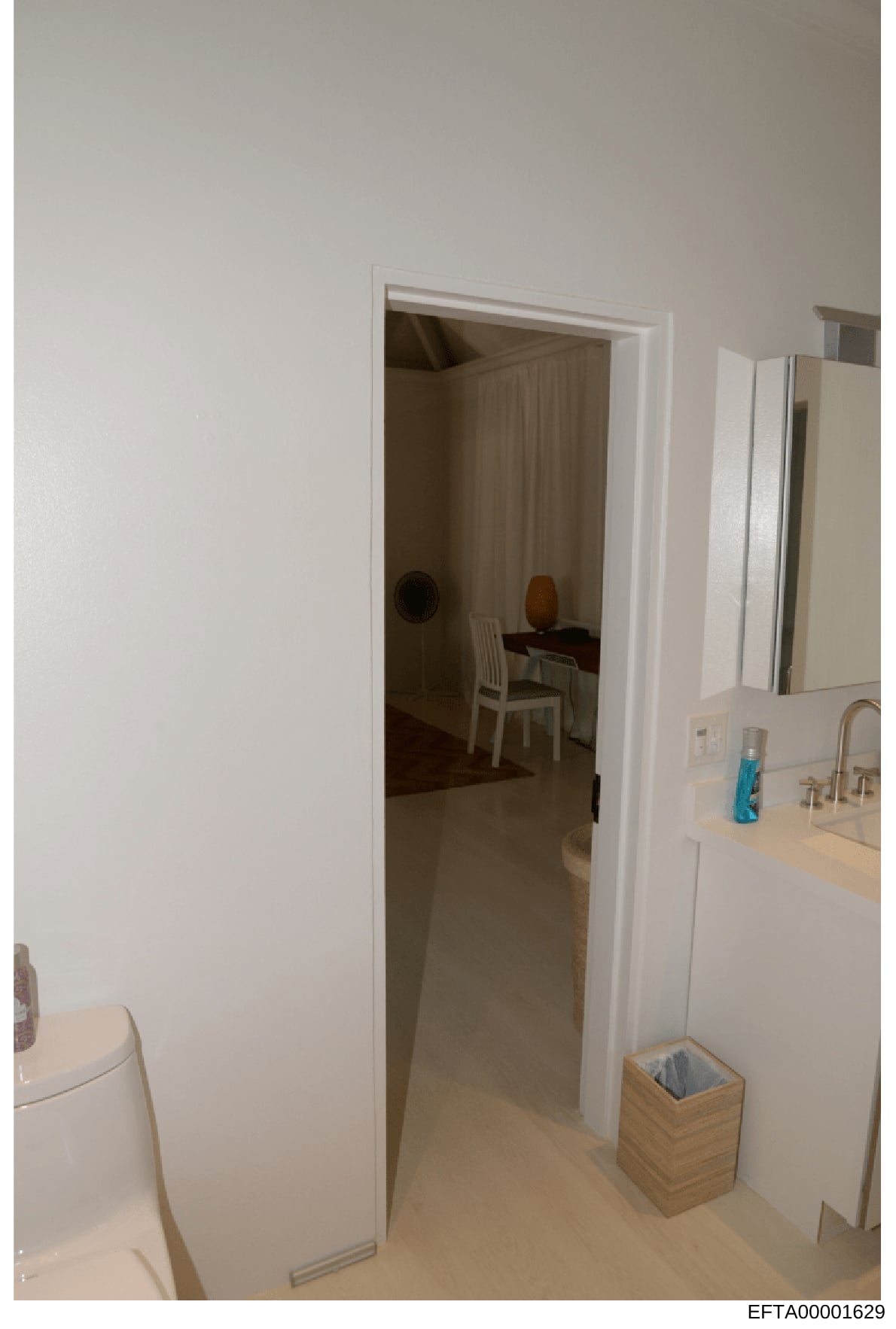 This photograph shows the interior of a residential bathroom with an open doorway revealing a bedroom beyond. The image appears to be evidence documentation from a property inspection or seizure, showing basic furnishings and room layout. The Bates n