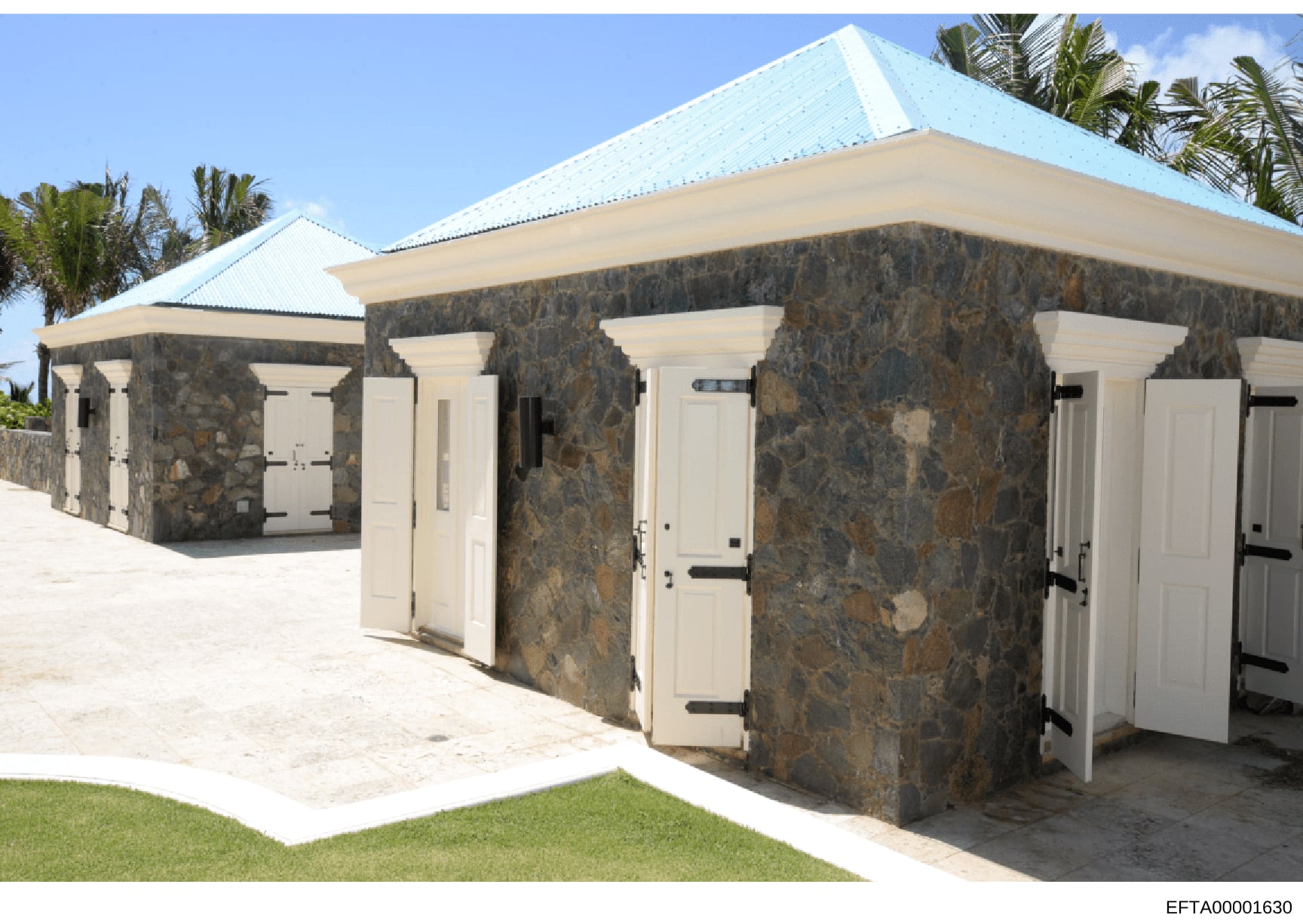 This is a photograph of a distinctive stone structure featuring multiple individual entrance doors, a bright blue metal roof, and cream-colored door frames. The building appears to be a changing room, bathhouse, or similar facility located on a tropi