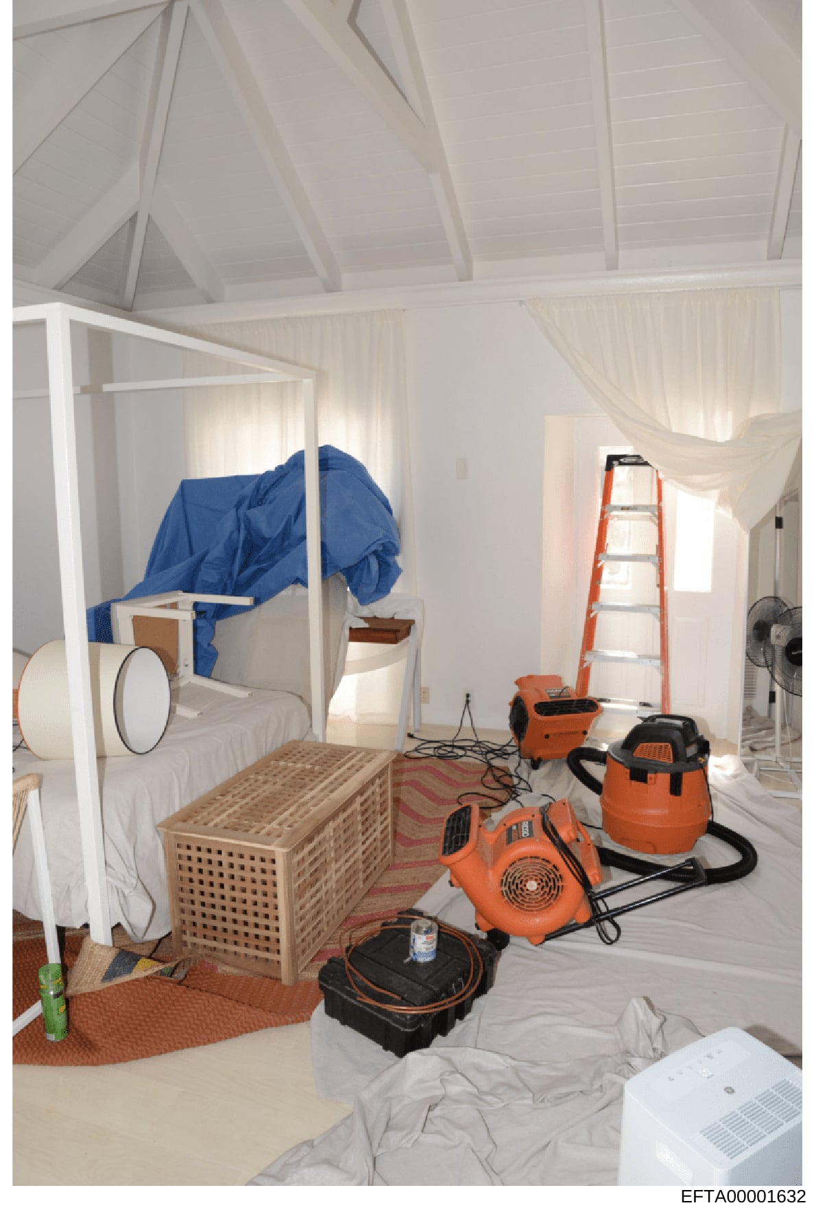 This photograph documents a bedroom undergoing water damage restoration or remediation work. The room contains multiple industrial drying and dehumidification machines, including orange air movers and a wet/dry vacuum, along with a ladder and blue ta