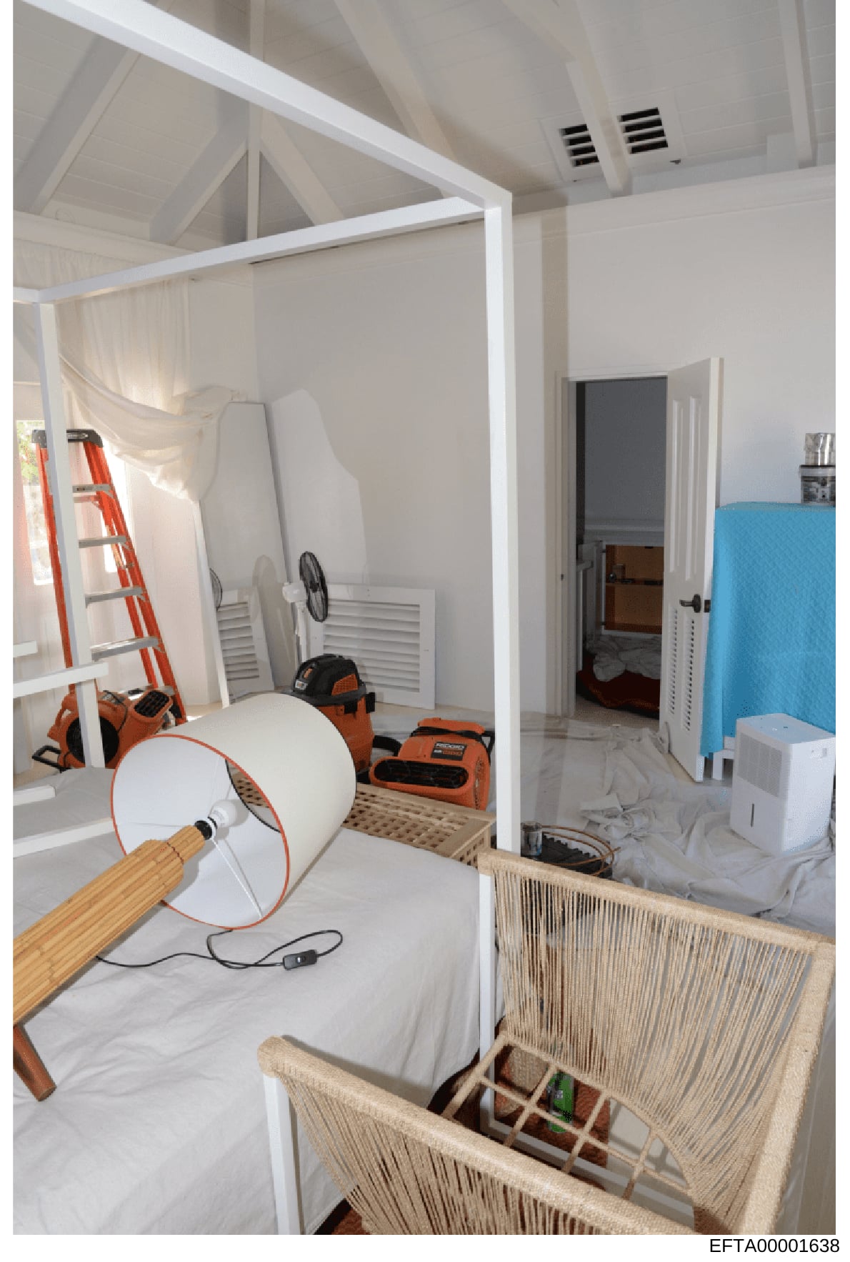 This photograph documents the interior of a room that appears to be undergoing renovation or construction work. The space contains various work equipment including ladders, fans, cleaning supplies, and power tools. The room features high ceilings wit