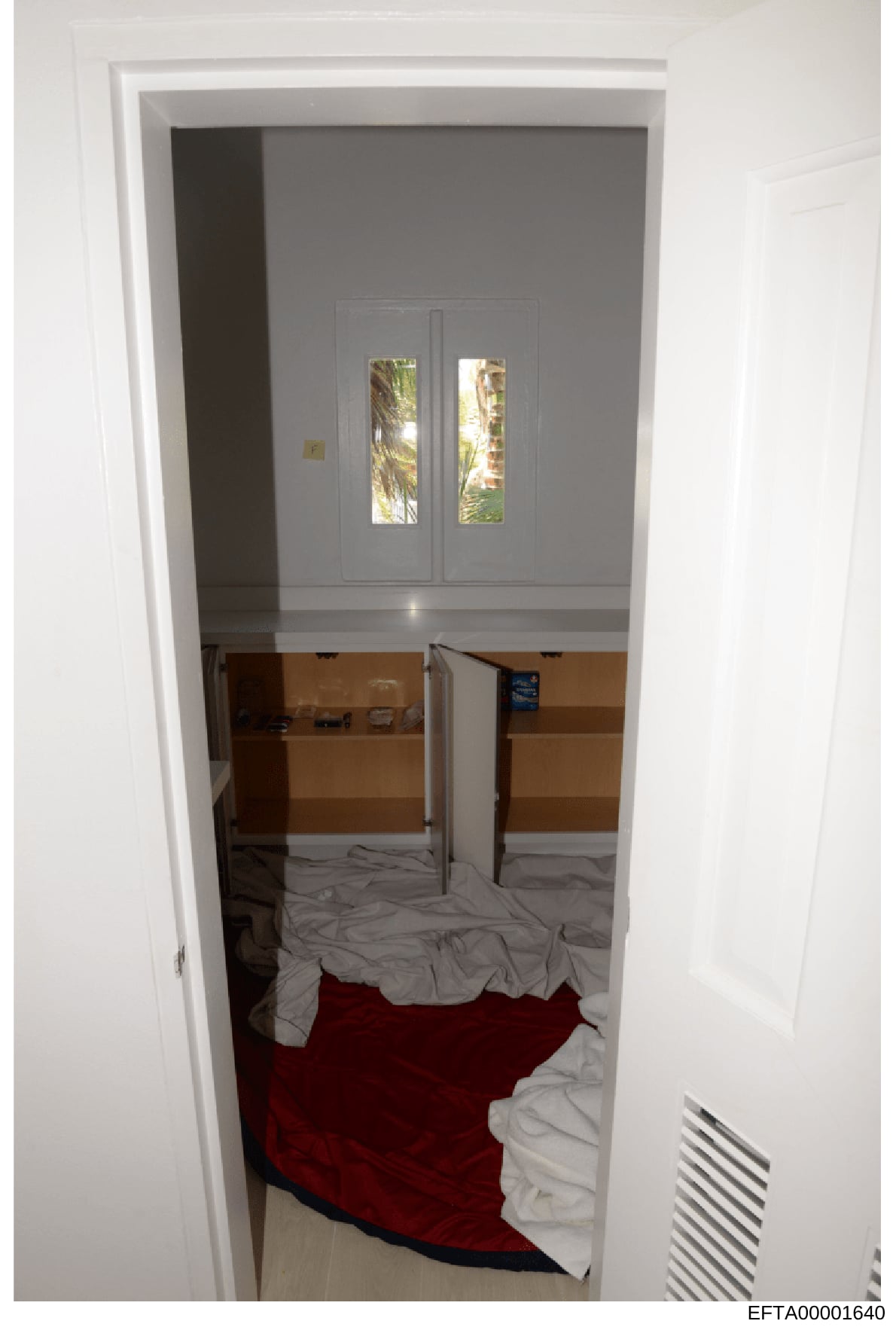 This is an evidence photograph showing the interior of a bedroom, taken from the doorway looking inward. The image depicts double doors with glass panes opening to an exterior area with tropical vegetation visible, wooden built-in cabinetry or shelvi