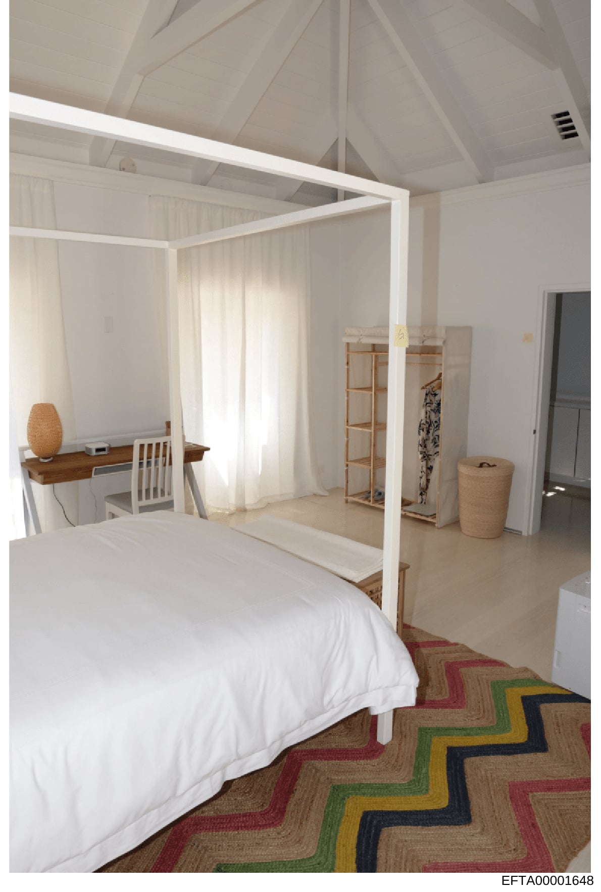 This photograph shows a spacious, minimally furnished bedroom featuring a white four-poster canopy bed with flowing curtains, a wooden desk with chair, exposed ceiling beams, and geometric patterned flooring. The room appears to be part of a property