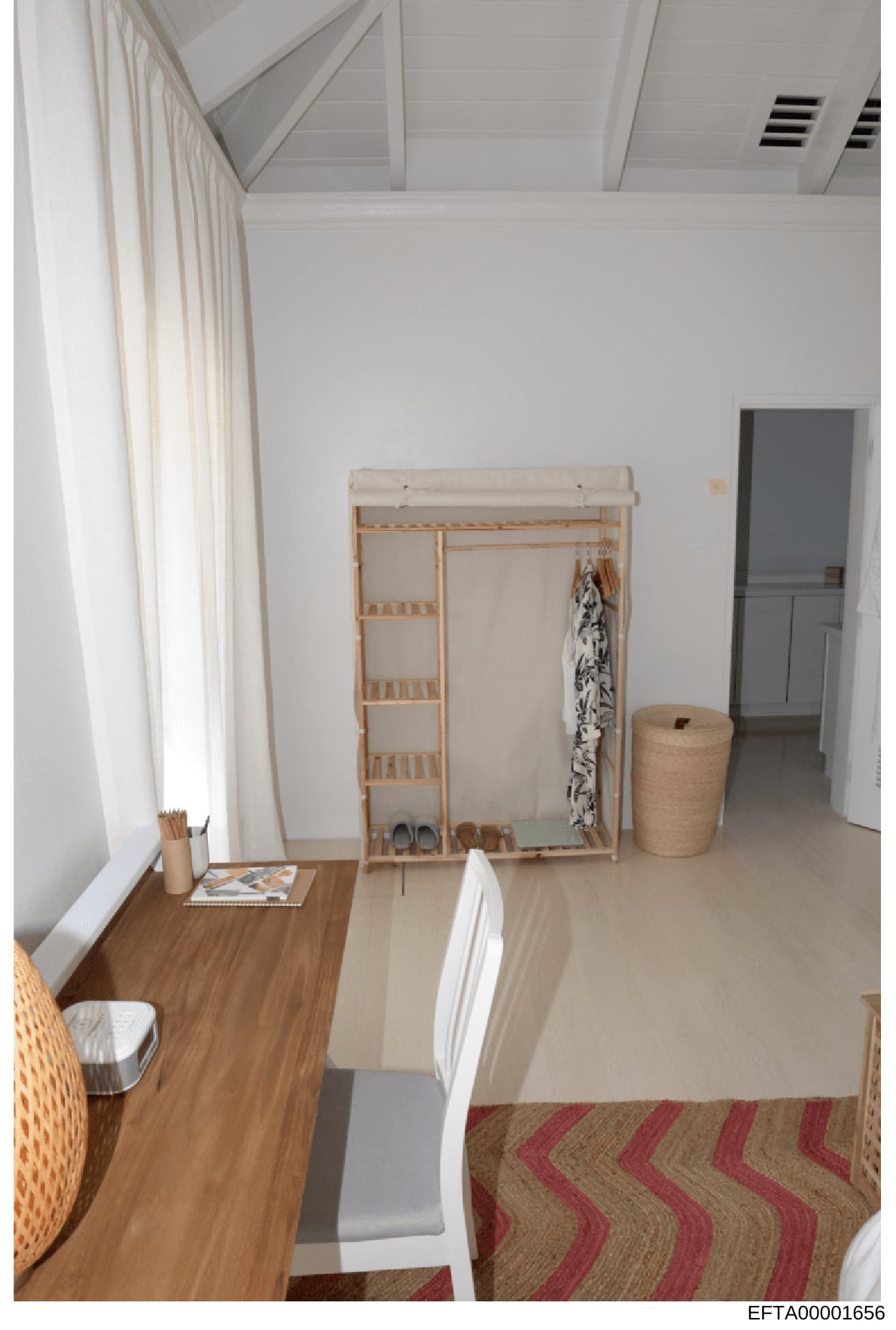 This is an overhead photograph of a sparsely furnished room with white walls, wood flooring, and minimal furnishings. The room contains a wooden desk with writing implements, a wooden wardrobe/clothing rack with hangers and garments, a woven storage 