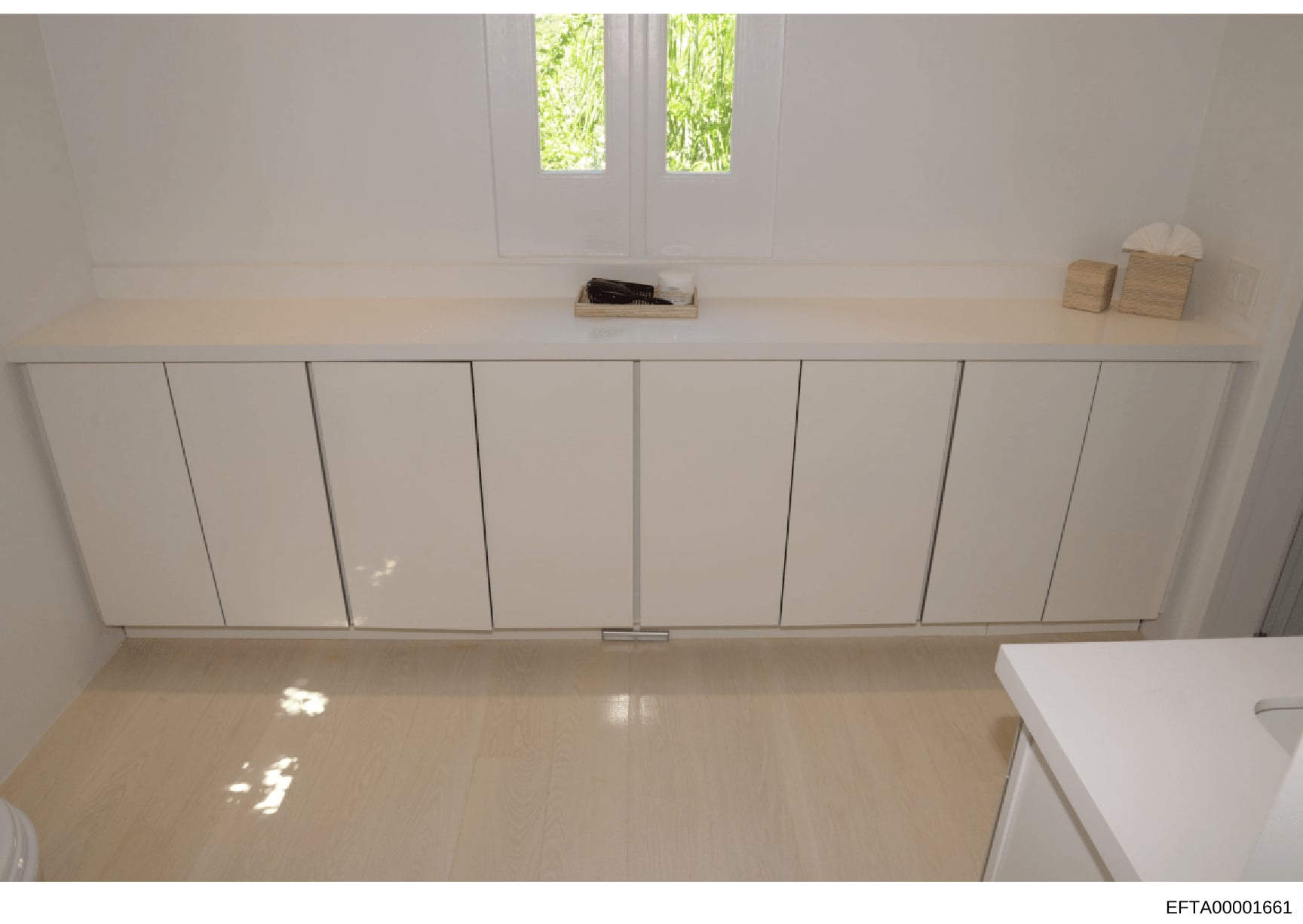 This is a photograph of a minimalist interior space showing a room with white built-in wall cabinetry, light wood flooring, and windows with natural light. The room contains minimal furnishings and appears to be part of a property documentation or ev