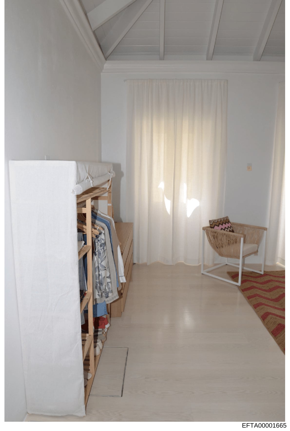 This is a photograph of a bright, minimalist interior space featuring a bedroom or dressing area with white walls, exposed ceiling beams, natural light from floor-to-ceiling curtains, and modest furnishings including a wooden clothing rack and wicker