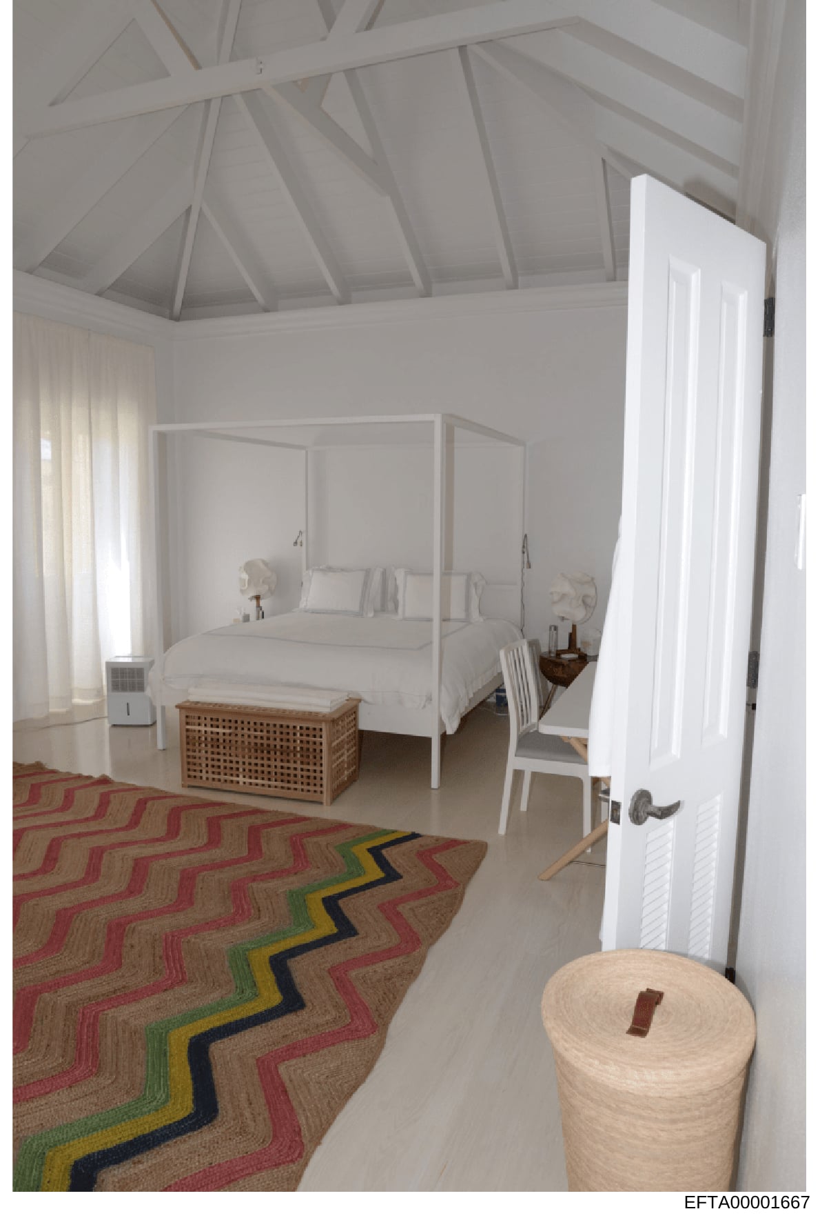 This is a photograph of an elegantly designed bedroom featuring a white canopy bed, vaulted ceiling with exposed beams, and contemporary furnishings. The room showcases high-end interior design with neutral tones, large windows with natural light, an