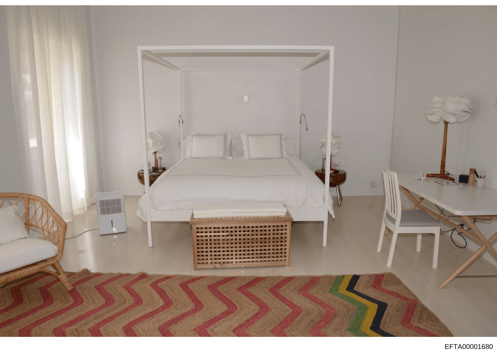 This is a photograph of a minimalist bedroom interior featuring a white four-poster bed with white linens, wooden nightstands, and basic furnishings. The room appears to be part of a property documented in the investigation, with sparse decoration an