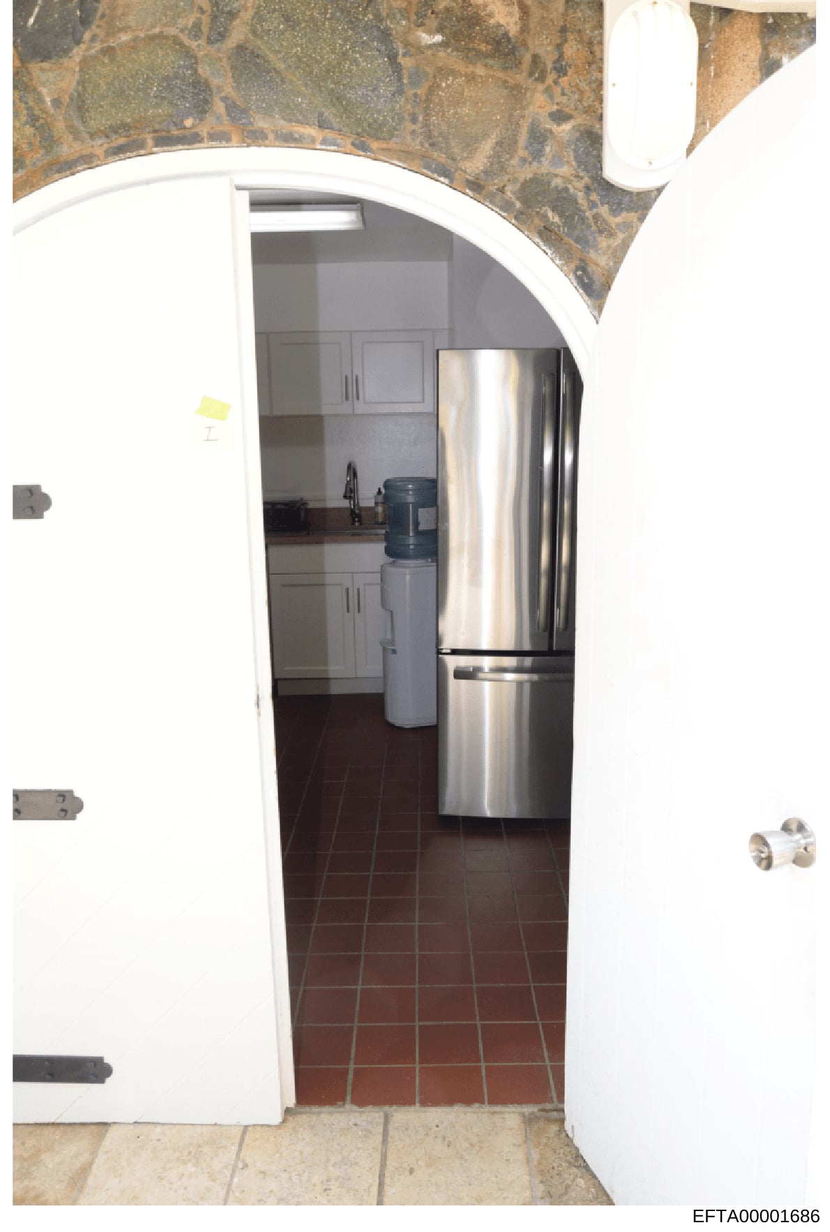 This is a photograph of a residential kitchen interior, taken through an open doorway. The image shows stainless steel refrigerator and appliances, white cabinetry, dark tile flooring, and decorative stone or marble wall accents. The photo appears to