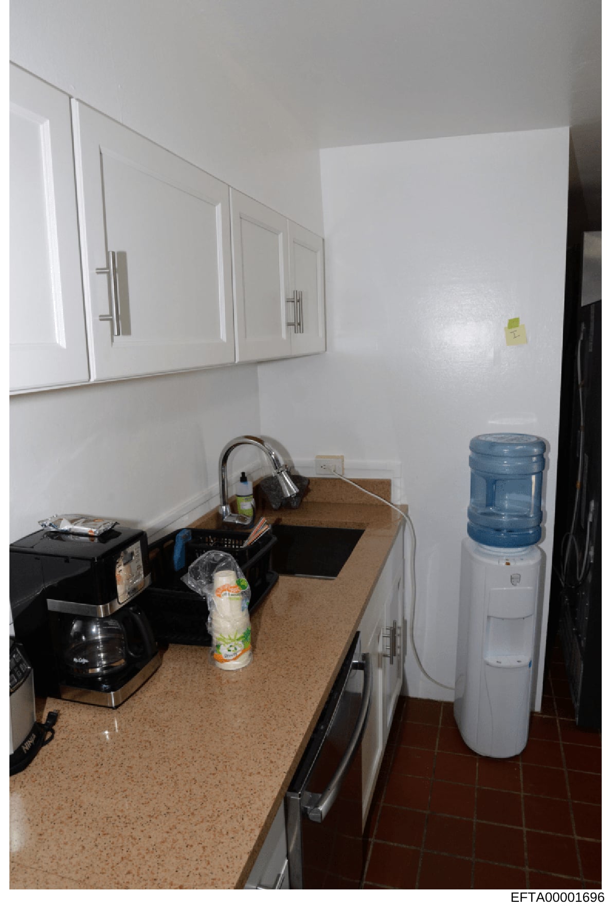 This is a photograph of a kitchen interior showing white wall cabinets, a beige countertop with various appliances including a coffee maker and water cooler, and basic kitchen fixtures. The image appears to be documentary evidence photography, likely