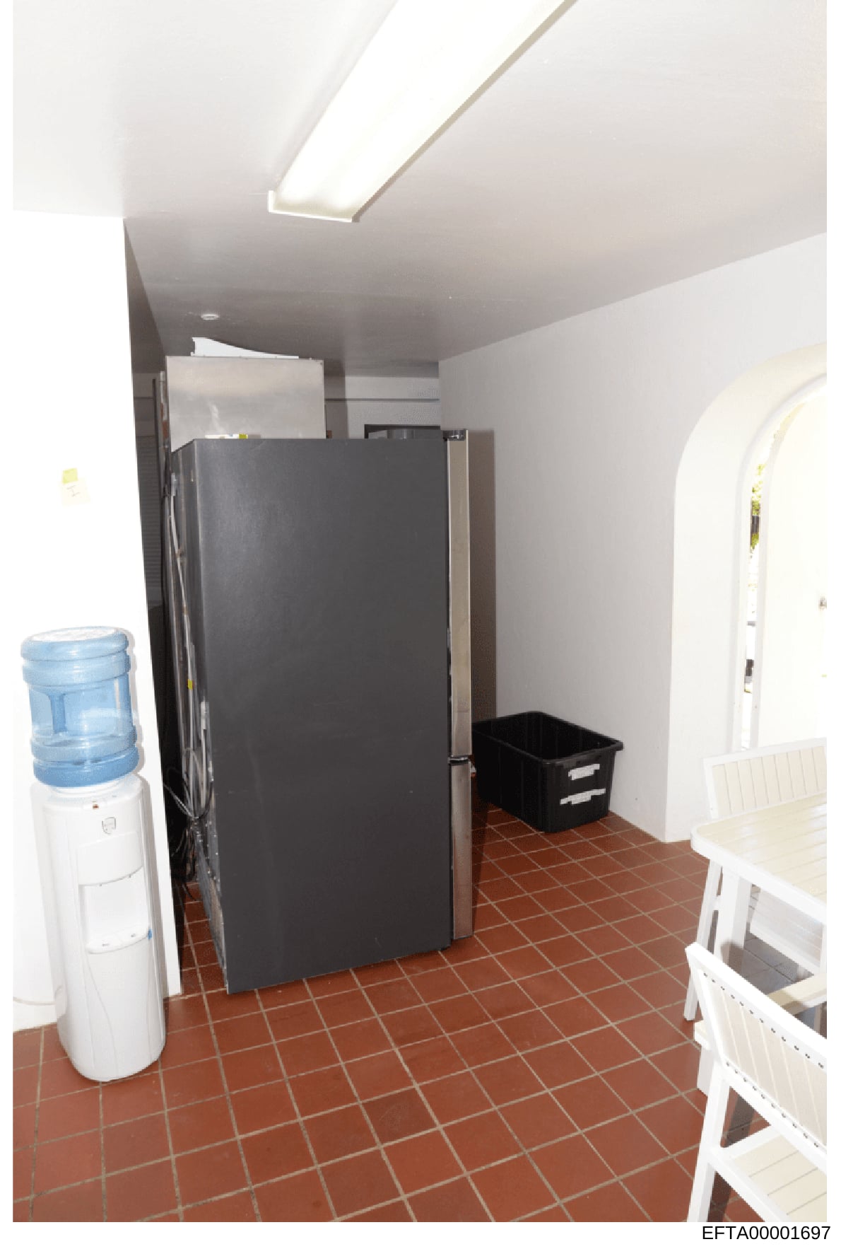 This is a color photograph of an interior room, likely a basement or utility area, showing a water cooler, refrigerator, black storage container, and white furniture or fixtures. The red-brown tile flooring and white walls with skylighting are visibl