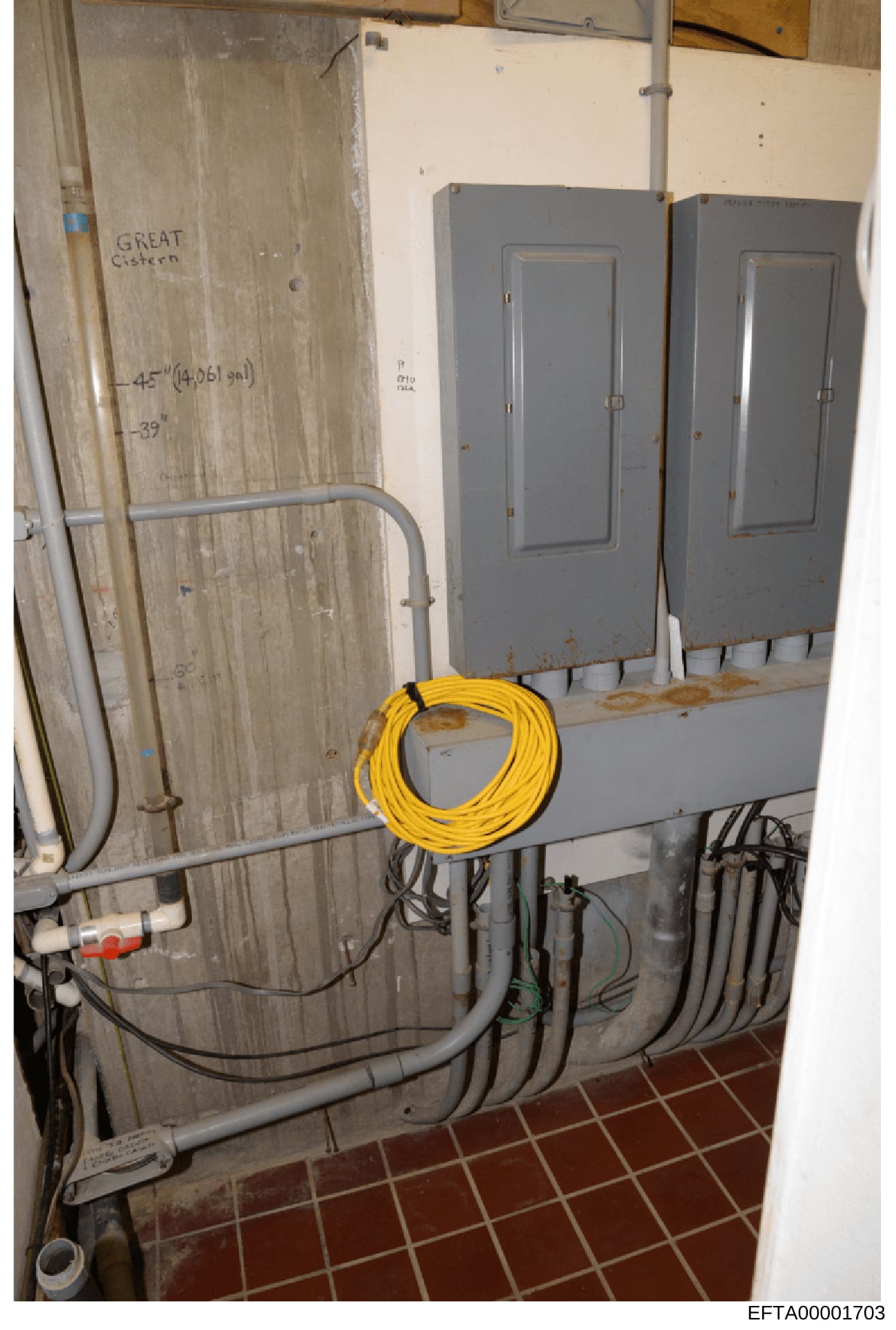 This is an evidence photograph showing electrical infrastructure in what appears to be a basement or utility area of an Epstein property. The image documents two gray electrical panels, extensive wiring, a coiled yellow extension cord, and handwritte