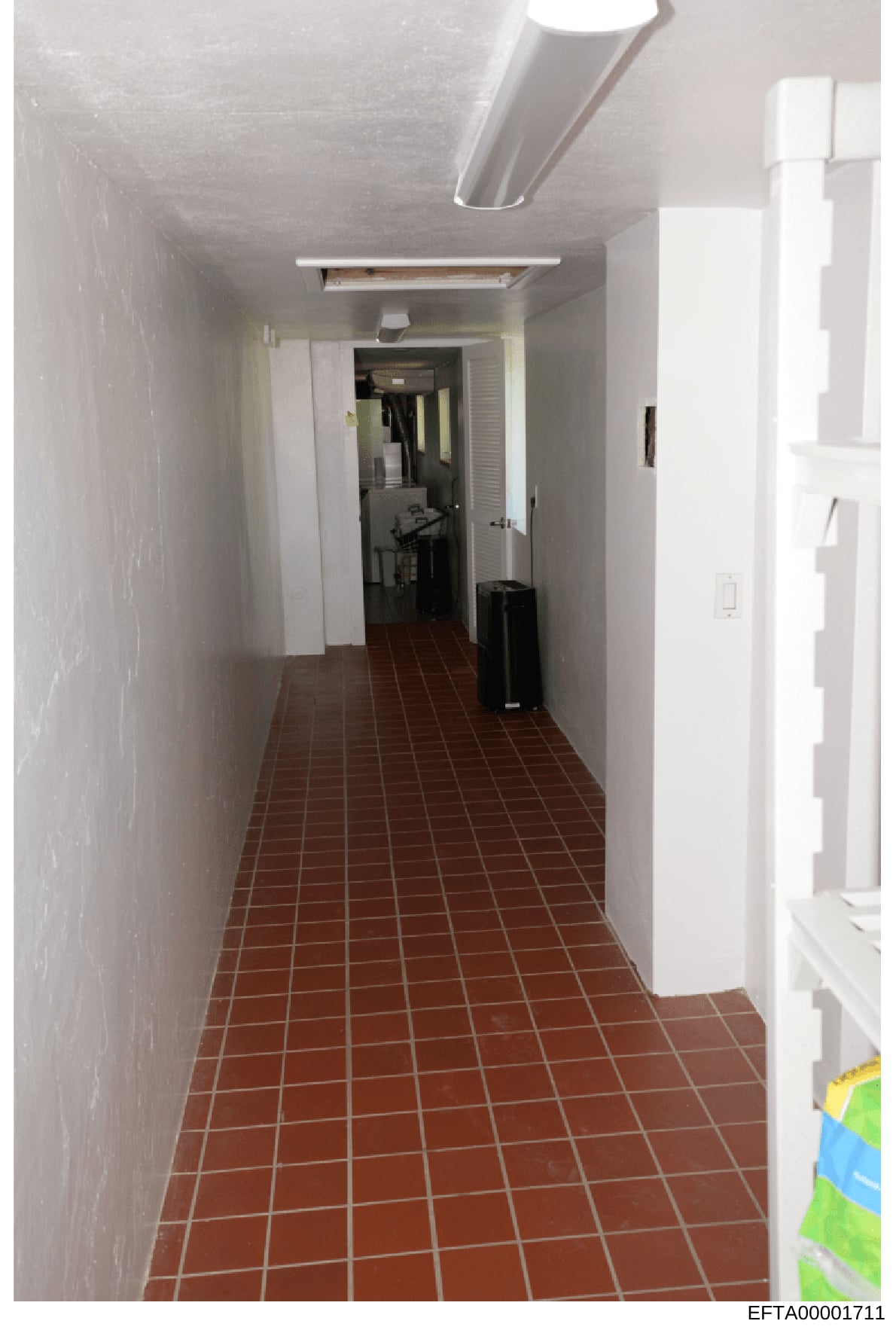 This is a photograph showing a long, narrow hallway with red/brown tile flooring and white walls. The hallway contains several doors on both sides, fluorescent lighting, and appears to be part of a commercial or residential building. The image is lik